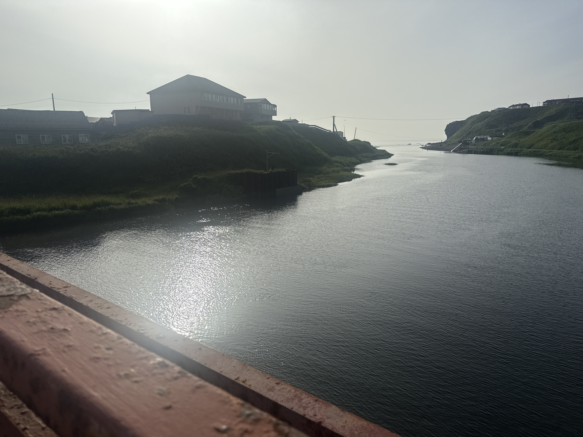 This is a photograph of a body of water, likely a river or bay, in Kuril'sk, Russia. The water is dark and reflects the overcast sky, with a bright sun flare visible. On the left side of the frame, a grassy hill slopes down towards the water, with several buildings situated on top. These buildings appear to be houses and possibly a larger institutional building, constructed in a functional, mid-20th century style. The hill is covered in dense, green vegetation. On the right side of the frame, the land rises into a steeper, grassy cliff that leads to the sea. More buildings are visible scattered along the top of this cliff. The foreground of the image shows a portion of a reddish-brown structure, possibly a bridge railing, which is out of focus. The overall impression is a coastal settlement under a muted sky, likely taken from a bridge or elevated vantage point overlooking the water.