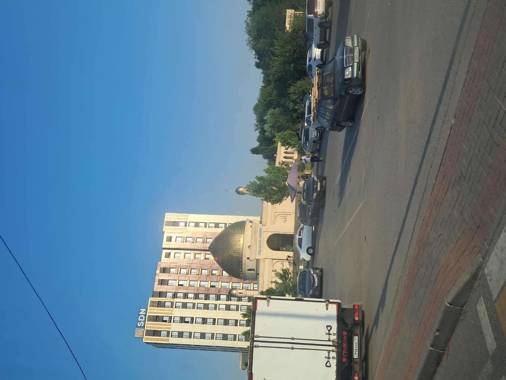 This media file depicts an outdoor scene with a clear blue sky, suggesting fair weather during the day. The image is oriented vertically, with the left side of the frame showing the sky and the right side featuring a street with parked cars, a building with a golden dome, and a tall apartment building.
The main subjects include several parked vehicles, including a white truck with a cargo box at the forefront, a dark-colored sedan with wooden planks on its roof, and other cars further down the street. A few people are visible near the building with the golden dome, some appearing to be walking. The building with the golden dome appears to be a mosque, indicated by its architectural style and the Arabic calligraphy visible on its facade. Adjacent to the mosque is a tall, modern apartment building with numerous windows. A sign on the apartment building reads "SDN" with a stylized logo. The street is paved with asphalt and bordered by a brick sidewalk.
The scene suggests a typical day in a city, possibly during a period of moderate traffic and pedestrian activity. The lighting indicates it is daytime, likely morning or afternoon due to the bright sky. The presence of a mosque and apartment buildings suggests an urban or suburban setting. The overall mood appears neutral, focusing on documenting the location and its elements.