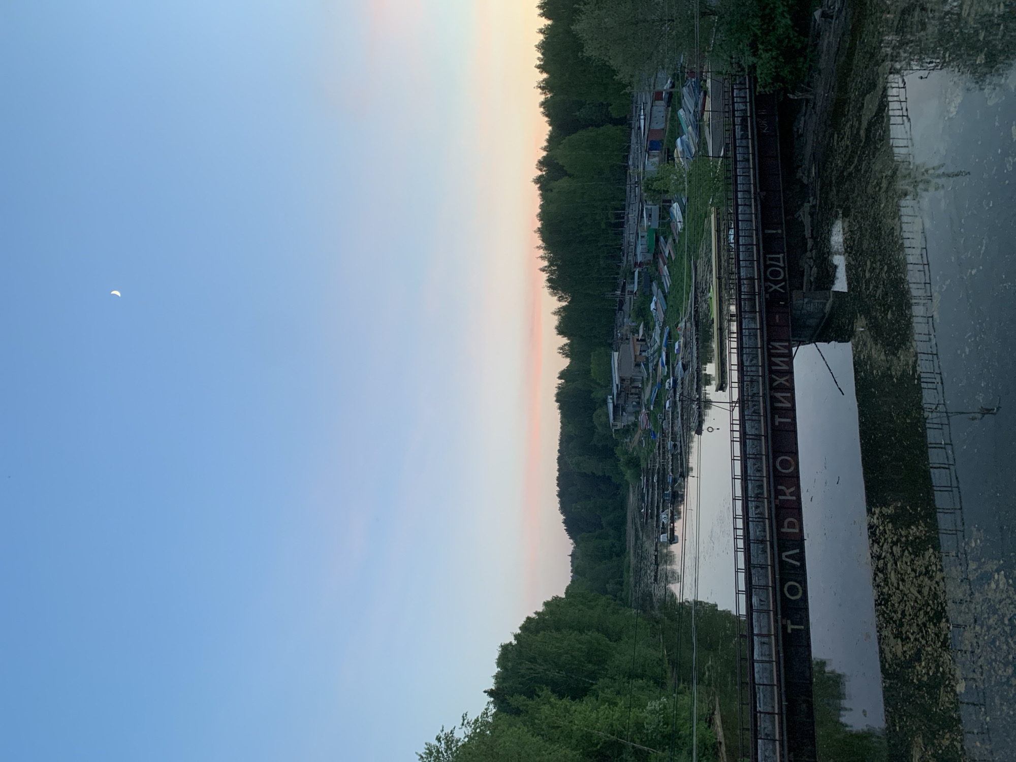 The image depicts a waterfront area in Chaikovsky, Russia, at dusk. A river is present in the foreground, with a metal walkway crossing over the water. The walkway has Cyrillic writing on its side. Along the riverbank, there are a series of small boat garages or sheds, which are clustered together. Dense vegetation, likely trees, lines the banks of the river. The sky is visible above, transitioning from a light blue to a pale orange-pink hue near the horizon. A crescent moon is visible in the sky. The water surface reflects the sky and surrounding structures.