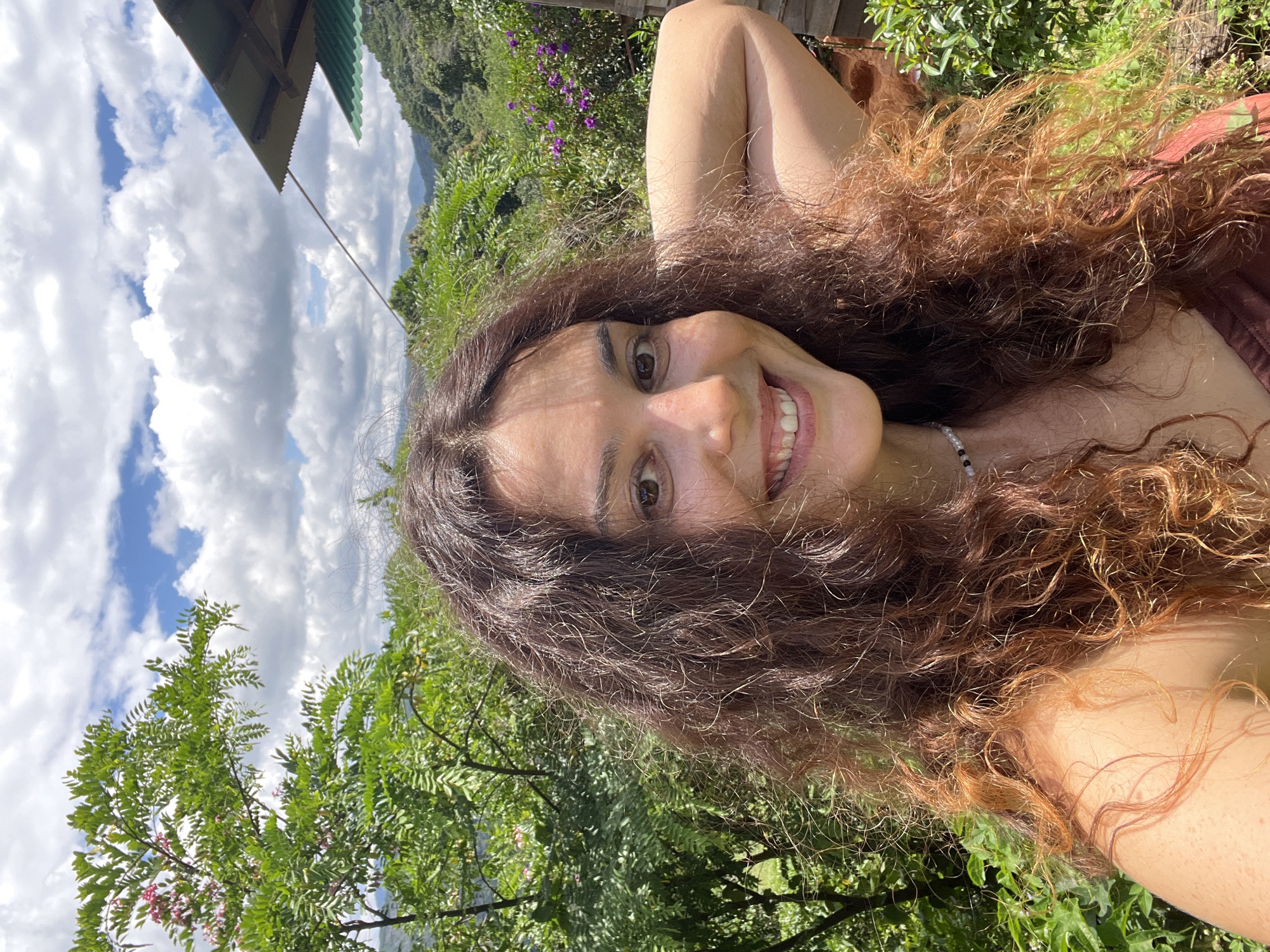 In a medium close-up selfie taken outdoors, a young woman with long, wavy brown hair smiles at the camera. She is wearing a brown top and a beaded necklace. Her right arm is bent behind her head, and her left arm is not fully visible. The background features lush green foliage, including a tree with pink flowers on the left, and rolling hills with a structure in the distance. The sky above is a bright blue with scattered white clouds. The scene suggests a natural, possibly rural setting. This location is identified as Sơn Điền, Vietnam.