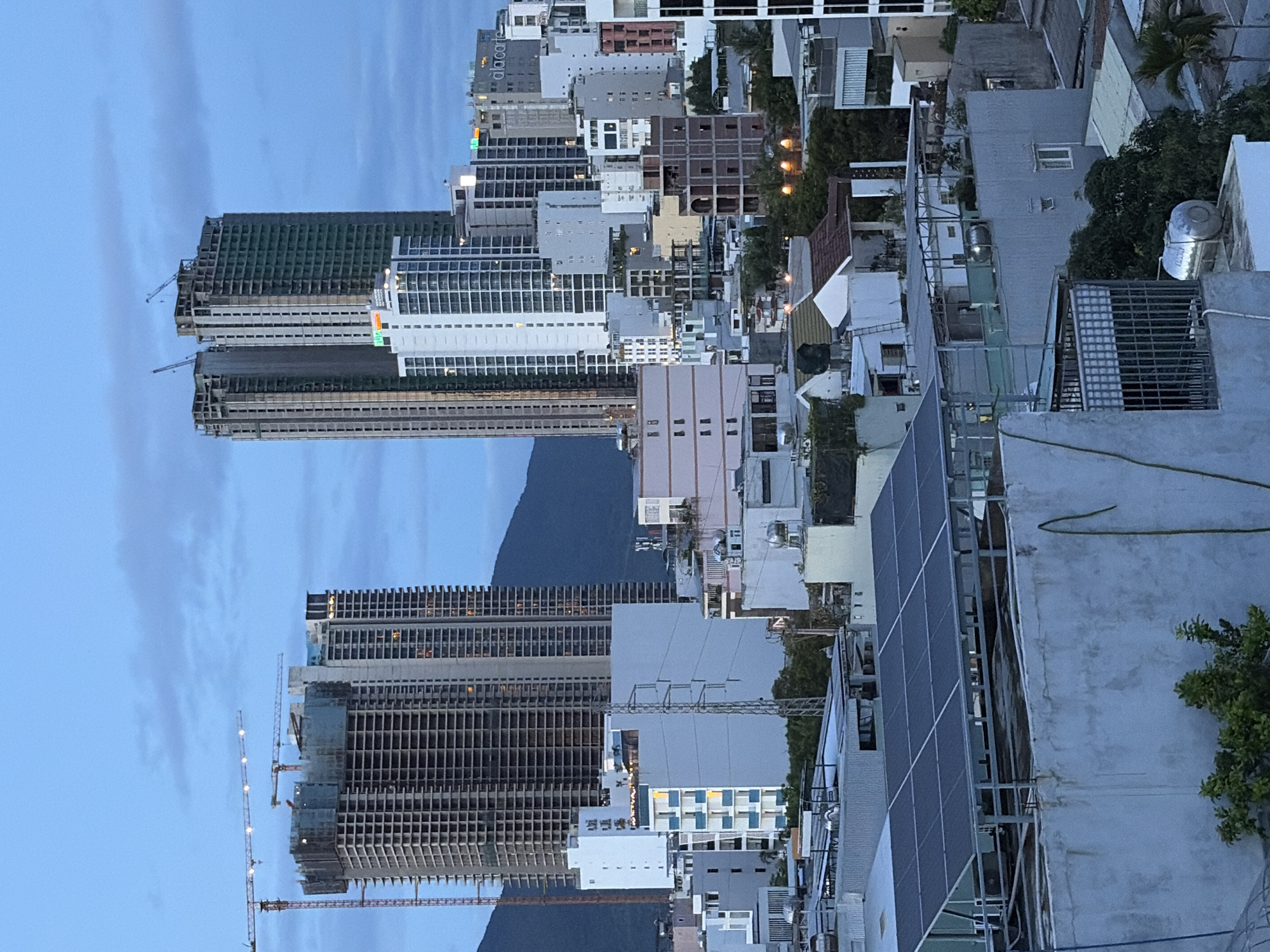 The image depicts a cityscape at dusk in Da Nang, Vietnam, characterized by a cluster of tall, modern buildings, many of which appear to be under construction. In the foreground, a rooftop is visible with a large array of solar panels. Several residential buildings and smaller structures are scattered throughout the mid-ground, with some showing illuminated windows, suggesting occupancy. In the background, mountainous terrain is faintly visible against a pale blue, cloudy sky. Construction cranes are prominent elements in the scene, particularly near the taller buildings, signifying ongoing development. The overall atmosphere is one of urban growth and transition.