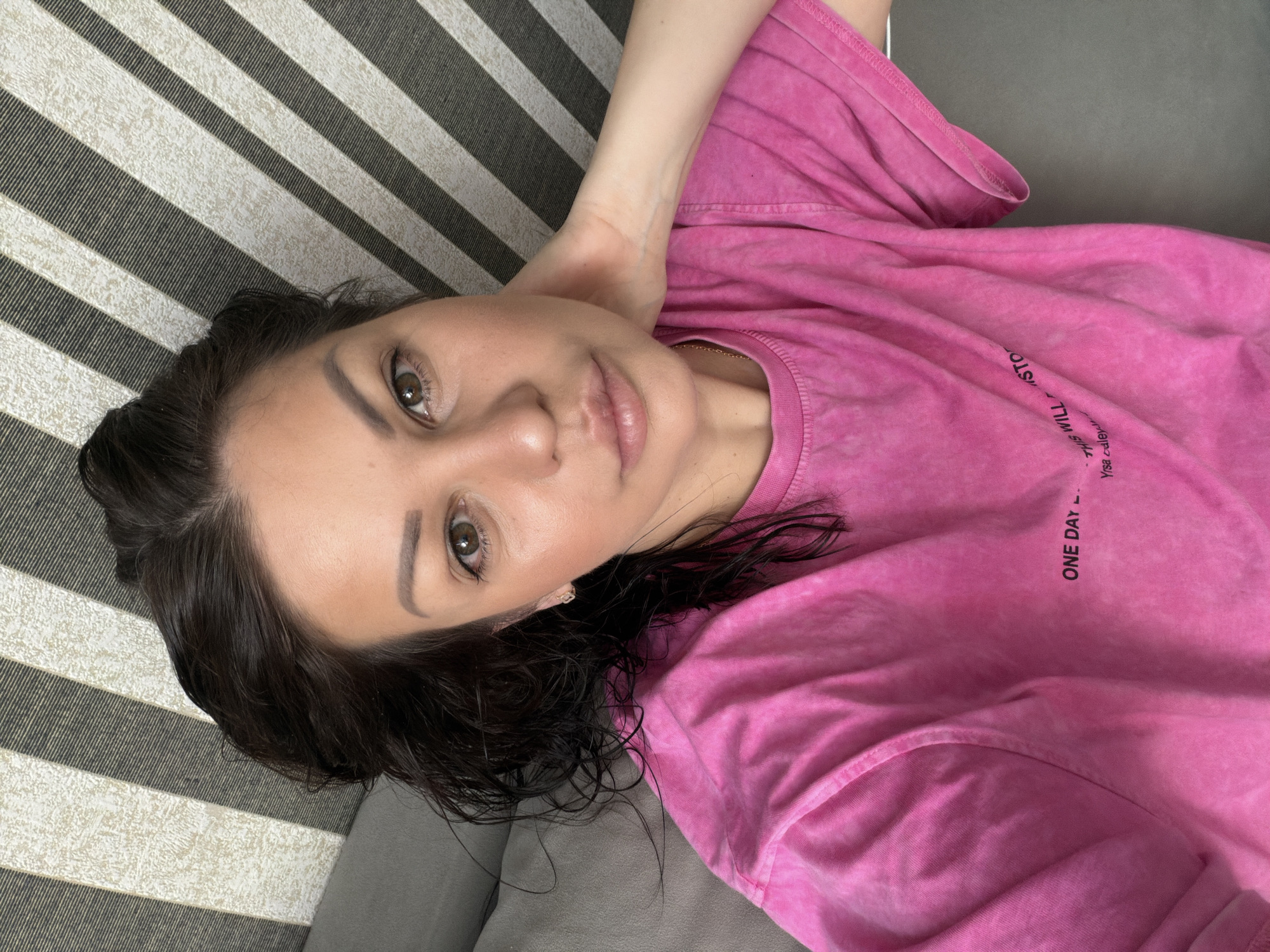 A woman is lying down, looking at the camera, with her hand behind her neck. She has dark hair and is wearing a pink t-shirt with text on it. The background appears to be a striped surface and a grey couch. The location is not specified as Sochi, Russia.