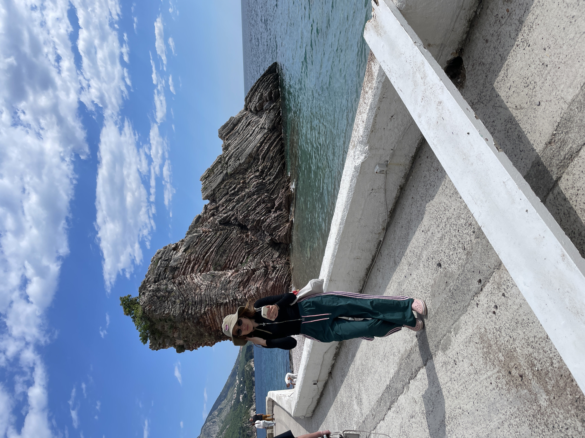 A medium shot shows a woman posing with a mobile phone on a concrete pier in Rafailovići, Montenegro, against a backdrop of a striking rock formation, a calm sea, and a partly cloudy sky.
The woman is wearing a black long-sleeve shirt, green track pants with pink stripes, pink shoes, a straw hat, and sunglasses. She is holding a mobile phone in her right hand and appears to be taking a photo or video. A handbag is hanging on her shoulder.
Behind her is a rocky island or peninsula rising from the sea, with trees growing on the top. The sea is calm and turquoise-green, with the sky blue with fluffy white clouds above.
The concrete pier is grey and has white concrete barriers. A small dark hole is visible on the pier close to the barriers.
The photo is taken in daylight, and the scene is well-lit. The presence of people in the far background indicates that this is a tourist area.