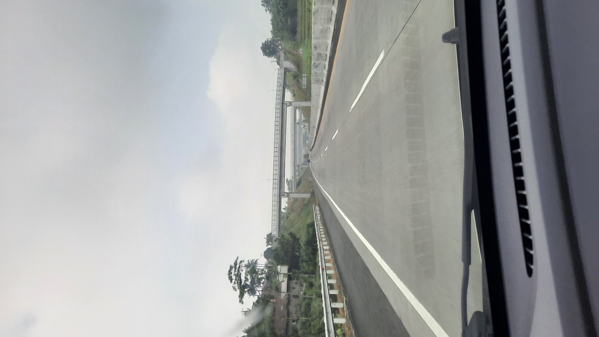 This is a photograph taken from inside a vehicle, looking out through the windshield at a highway. The highway stretches into the distance, with multiple lanes separated by white lines. There are overhead bridges crossing the highway, and a white guardrail runs along the side of the road. The landscape is hilly and green, with trees and vegetation visible. In the foreground, the dashboard of the vehicle is visible, including the windshield wipers. The sky is overcast and the overall lighting suggests it is daytime, possibly early morning or late afternoon. The location is identified as the city of Ngampon, Indonesia. There are no people or animals visible in the image. The scene is one of transportation and infrastructure in a semi-rural setting.