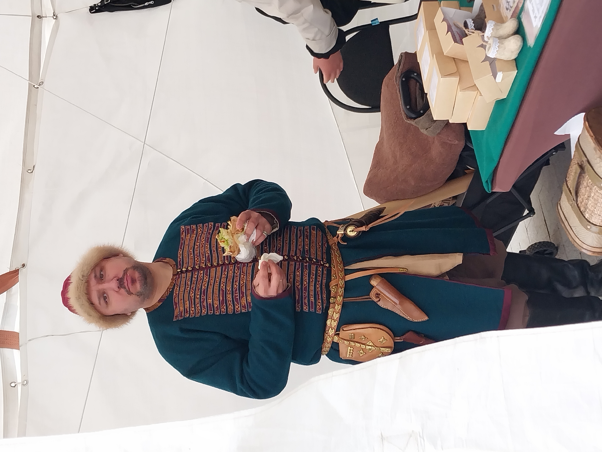 In Orenburg, Russia, a man dressed in a teal-colored traditional costume with intricate detailing around the collar and down the front is eating a snack. He wears a fur-trimmed hat and has a brown belt with a leather pouch and knife sheath. He is standing in a tent with a white canvas roof. To his right is a table covered with a green cloth, displaying small boxes and handmade shoes. A brown bag sits on the table, and a person wearing a white jacket stands next to it.