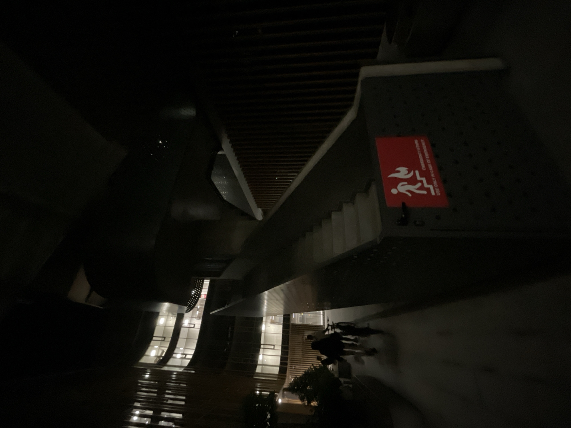This media file captures a nighttime scene in Sofia, Bulgaria, characterized by low light and a modern architectural setting. The perspective is angled downwards, offering a view of illuminated interior spaces within a building, possibly a shopping center or office complex. The architecture features curved walls and extensive glass facades, revealing multiple floors with bright interior lighting.
Prominently displayed on a dark, perforated surface is a red emergency exit sign with white pictograms. The sign depicts a figure running down stairs with flames on one side, indicating "EMERGENCY EXIT" and "USE ONLY IN CASE OF EMERGENCY." The texture of the surface suggests a metal grate or perforated paneling, possibly part of a ventilation system or decorative element.
In the foreground, partially obscured by shadows, are several figures. They appear to be walking, their forms silhouetted against the ambient light. The presence of these individuals suggests activity within the building or its immediate surroundings, despite the late hour. Reflections on a dark, polished surface below hint at the building's interior and possibly outdoor elements like trees, adding depth to the scene.
The overall mood is somewhat mysterious due to the low lighting and high contrast. The bright interiors juxtaposed with the dark exteriors create a sense of urban nighttime ambiance. The visible text on the emergency sign provides a functional detail within the aesthetic environment.