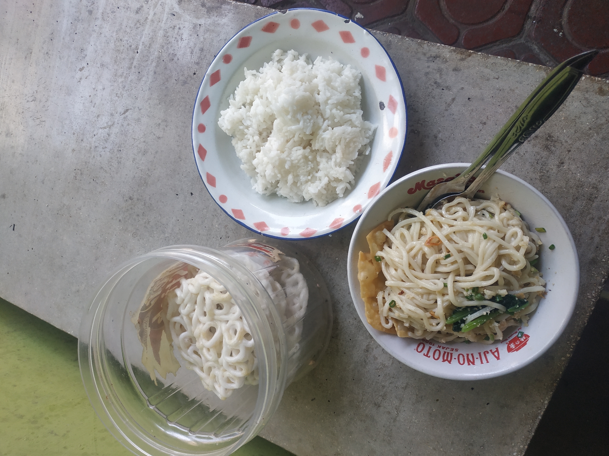 This image shows a table setting with food. In the foreground, a white bowl with red and blue trim contains a portion of noodles with some green vegetables mixed in, and a few pieces of fried wontons on top. The bowl has the words "AJI-NO-MOTO" and "SEJAK 1900" printed on it in red lettering. A spoon with a green handle rests on the edge of the bowl. To the right and slightly behind the noodle bowl is another white bowl with a similar red and blue trim, filled with a generous serving of white rice. To the left and further back, a clear plastic container is filled with white, circular crackers, possibly krupuk. The food is arranged on a textured gray concrete surface, with a hint of a green wall visible in the upper left corner. The background shows a reddish-brown tiled area. The overall impression is of a simple, home-style meal. The presence of the Ajinomoto branding suggests a possible location within Indonesia, and the specific phrase "SEJAK 1900" indicates that this is a product that has been around since that year. While the image itself does not explicitly place the scene in Jember, Indonesia, such a meal and the prominent display of Ajinomoto are common in Indonesian cuisine.