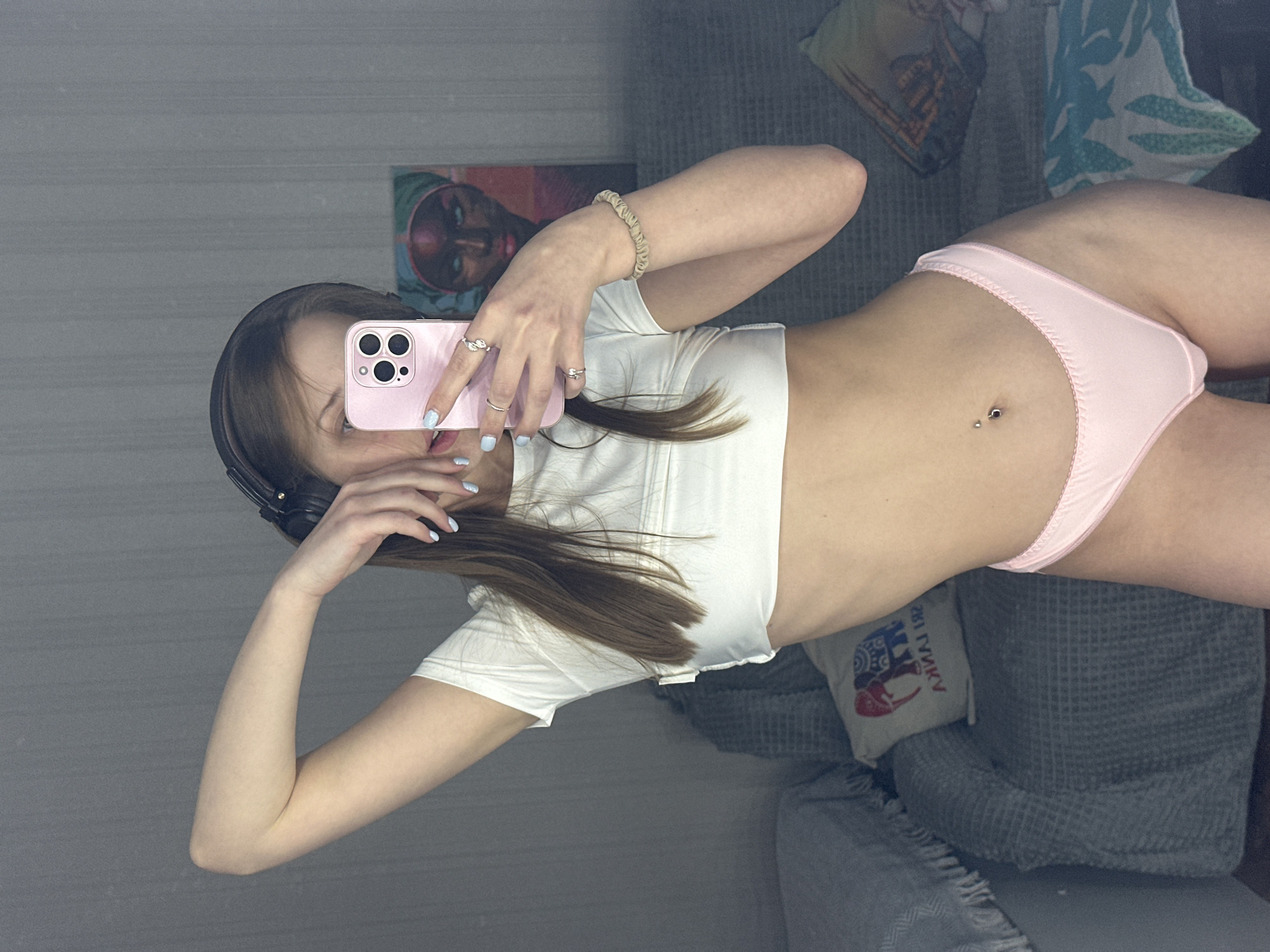 A young woman is taking a selfie in a mirror. She has long brown hair and is wearing a white crop top and pink underwear. She is also wearing headphones and several rings on her fingers. The background shows a couch with pillows and a painting on the wall. The room appears to be dimly lit. The woman's expression is neutral. There is a phone in her hand, reflecting the scene in the mirror.