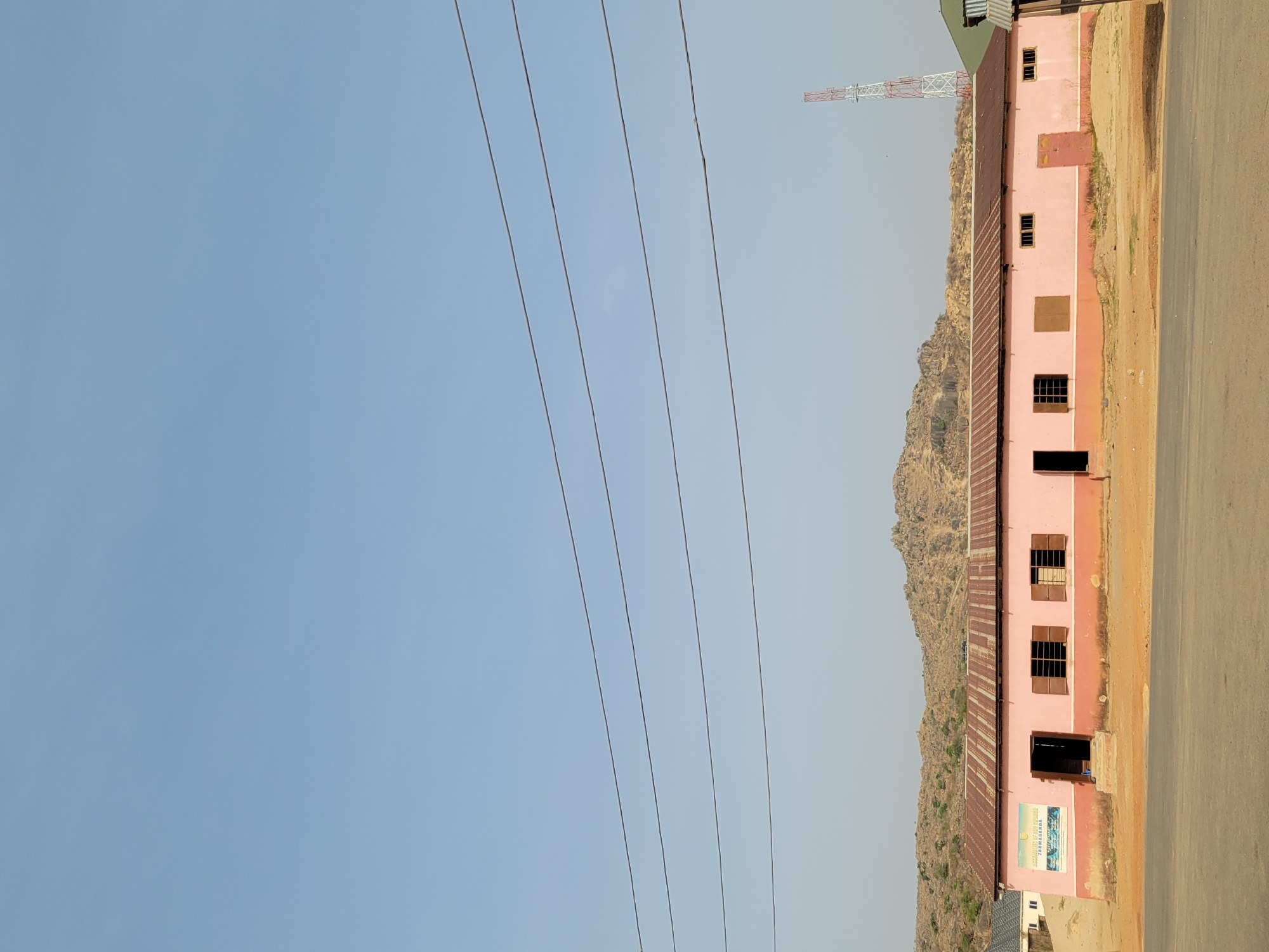 A slightly high-angle, horizontally-oriented outdoor daytime shot shows a pink, two-story building with a brown roof, five power lines running diagonally from left to right, and a communication tower in the upper right, in Gura Top, Nigeria. Behind the building, there are low, rocky mountains. The front of the building faces the viewer. The building has a series of rectangular windows, some with shutters, and one open door on the first floor. A sign is visible on the lower left corner of the building. To the right of the building is a stretch of road or open, dirt-covered ground. The sky is blue.