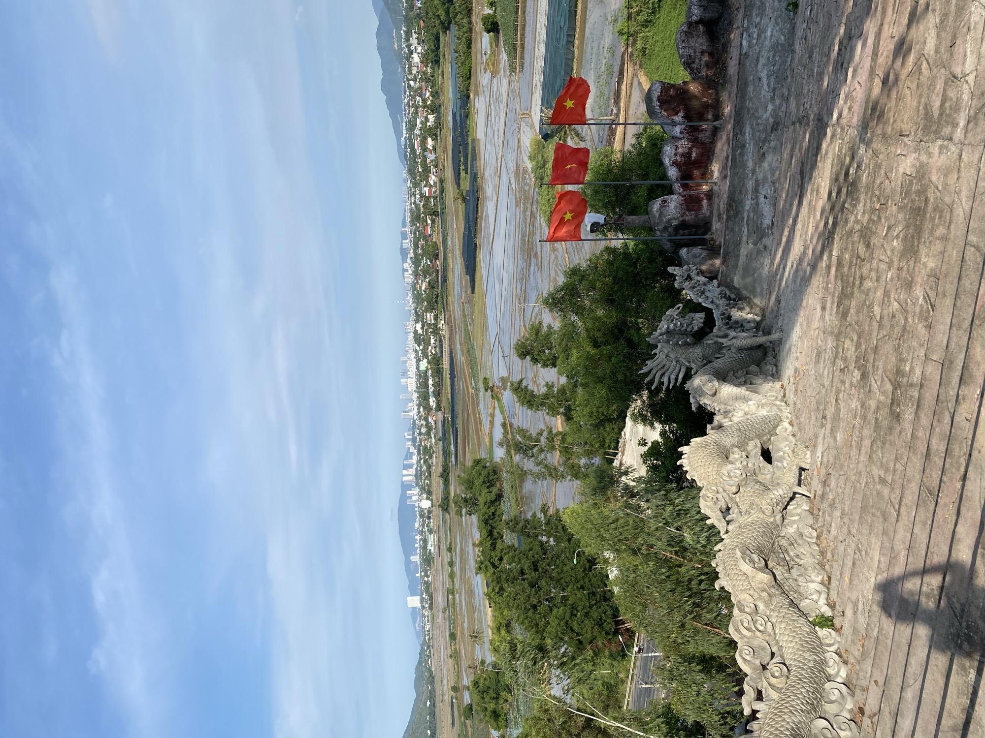 The image captures a scenic overlook in Tay Nha Trang Ward, Vietnam, offering a panoramic view of a city skyline nestled between rolling hills and expansive flooded rice paddies. In the foreground, a stone dragon railing, a common motif in Vietnamese architecture, lines a set of concrete stairs. Three Vietnamese flags are prominently displayed on poles to the right of the railing, fluttering in the breeze against the backdrop of the distant cityscape. The landscape is a mix of lush greenery, including trees and shrubs, interspersed with the reflective surfaces of the flooded fields, creating a mosaic of textures and colors. The sky above is a clear blue, with scattered white clouds, suggesting a bright and pleasant day. The overall impression is one of natural beauty blended with urban development.