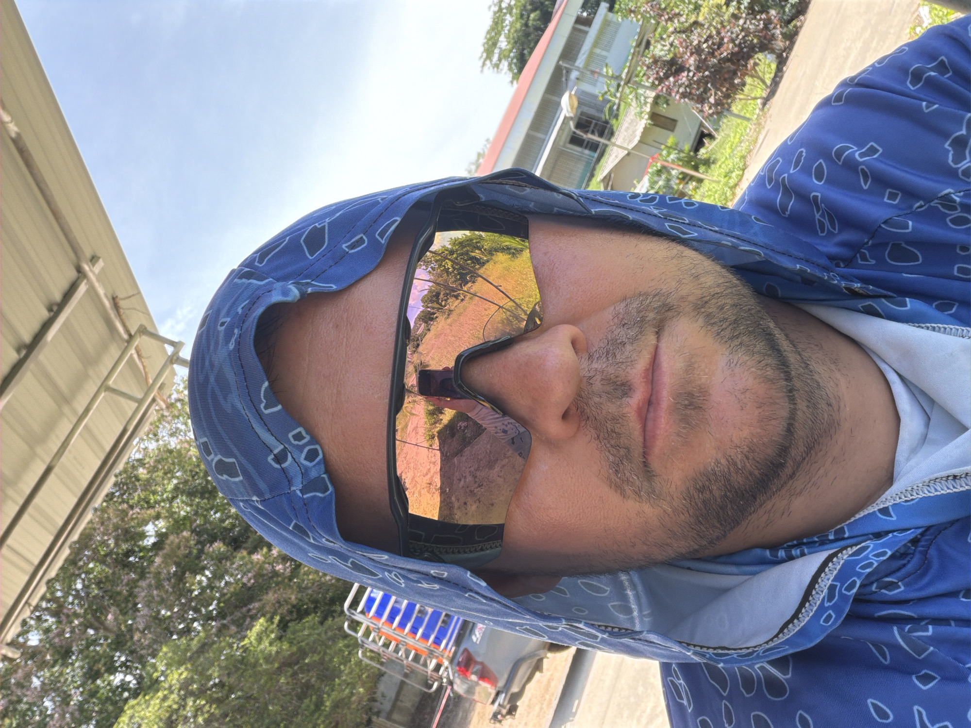 A close-up, selfie-style, medium shot captures a person wearing a blue hooded jacket with a white paisley-like pattern. The person has on oversized, reflective sunglasses that mirror the surrounding environment, showing a street, vegetation, and a building. The subject has stubble on their face and their hood is up, obscuring part of their head. In the background, to the left, a silver truck with blue crates on its rack is visible. To the right, a light-colored building with a red roof and a white porch is seen, surrounded by trees and greenery. The overall setting appears to be outdoors, possibly in a rural or semi-rural area of Baan Bo Phut, Thailand, given the architecture and vegetation. The lighting suggests it is daytime, with a bright sky.