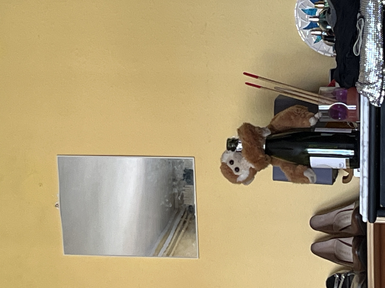 This image captures an indoor scene, likely a personalized corner within a room. The dominant background is a plain, warm yellow wall.
The central focus of the image is a small, light brown stuffed animal resembling a monkey, which appears to be playfully clinging to the neck of a dark green bottle. The bottle, possibly champagne or wine given its shape, features a white label whose text is not legible.
Behind the bottle, two slender sticks with red tips, resembling incense sticks, lean against a dark, flat surface, which seems to be a shelf. To the right of this arrangement, a higher shelf holds a decorative dish with blue and white mosaic-like pieces and a piece of shiny, sequined fabric or clothing. Below the shelf with the bottle, two pairs of women's dress shoes—one light brown and one darker brown, both heels—are neatly arranged.
To the left of the central display, a rectangular mirror is affixed to the yellow wall. The mirror's reflection is indistinct, showing a blurry indoor space with faint horizontal lines, possibly from window blinds or furniture.
The scene is static, depicting a collection of personal items rather than an activity or event. The time of day and weather are not discernible due to the indoor setting. While the location context is provided as Barcelona, Spain, there are no specific visual cues within the image itself to confirm this. No legible text is visible in the frame.