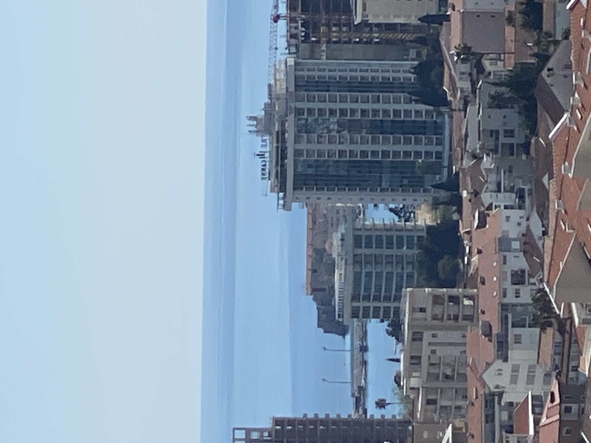 The image shows a cityscape in Budva, Montenegro, with a clear blue sky above the Adriatic Sea. Multiple high-rise buildings are under construction, including one with a visible crane. Completed buildings and residential houses with terracotta roofs are densely packed together. Roads, trees, and other urban elements are also visible.