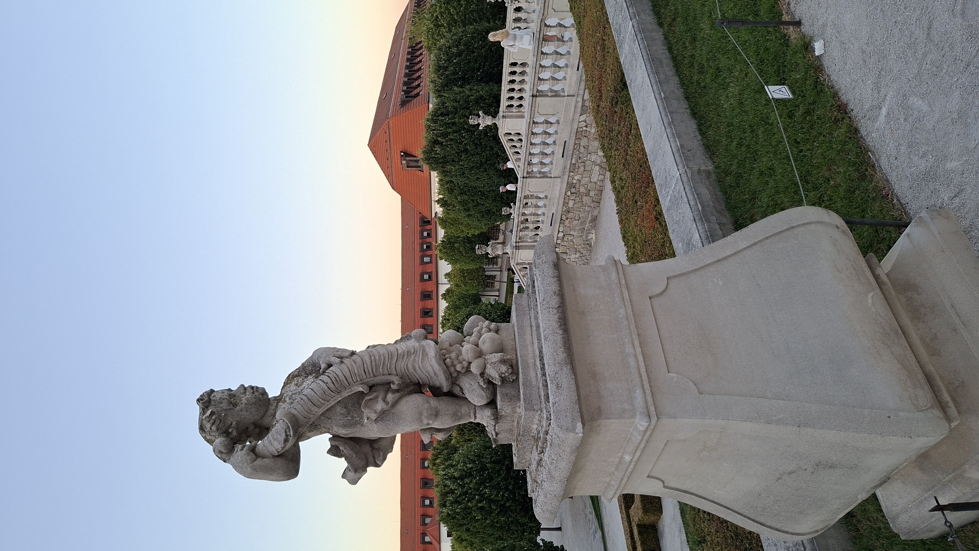 This is a photograph taken in Bratislava, Slovakia, likely during sunset, at Bratislava Castle. The foreground features a stone statue of a cherub or putto on a pedestal. The cherub is depicted as plump and is holding a garland of fruit. The pedestal is ornate, with a wide, curved base. Behind the statue, a series of white stone balustrades form a grand staircase leading up towards a large building with a red tiled roof. The building appears to be part of the castle complex. Green trees form a line behind the balustrades, and manicured hedges are visible in the mid-ground. The sky is a clear gradient of pale blue to orange, indicating the setting sun. A few people are visible on the staircase, one woman in a white dress standing on the upper section, and a few figures in the distance. The ground is paved with gravel.