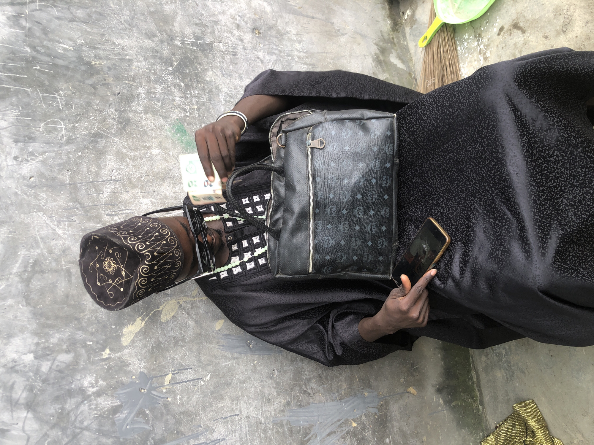 A man wearing a black traditional cap and black garment, adorned with a beaded necklace and sunglasses, sits leaning against a textured gray wall. He holds a wad of currency in his right hand and a mobile phone in his left. A black handbag with the "MCM" logo rests on his lap. The ground beneath him is concrete, with a broom and a green dustpan visible to the right. The scene appears to be an outdoor setting in Ado, Nigeria.