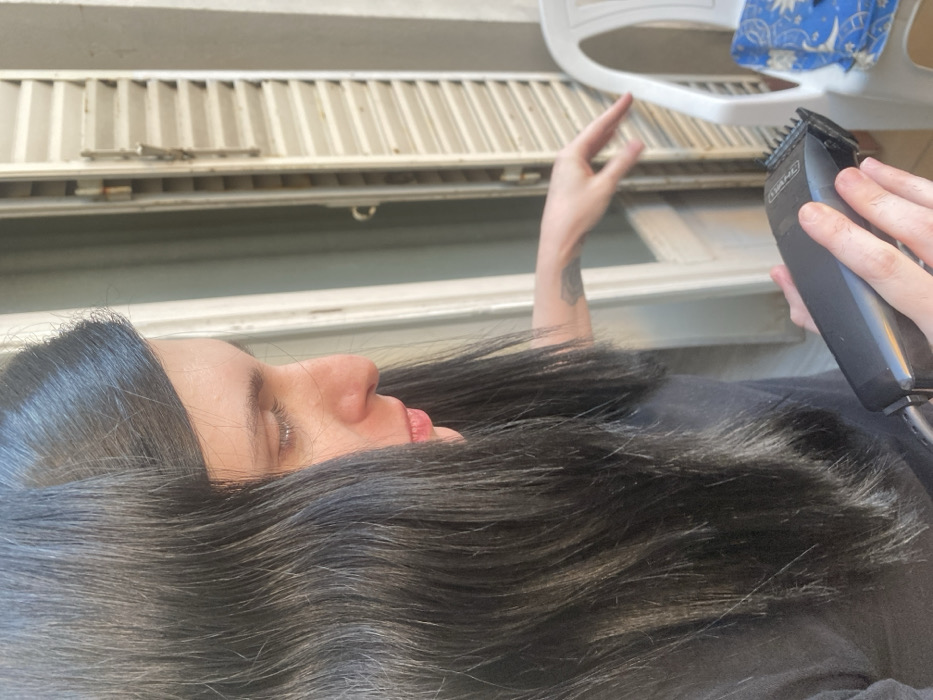 A woman with long, dark hair lies on her back. She has a tattoo on her left forearm. Her eyes are closed. Her hair is spread out around her face. In her right hand, she holds a black electric hair clipper. Above her, there's a white radiator and part of a white plastic chair with a blue and white patterned item resting on it. The background appears to be an open window.