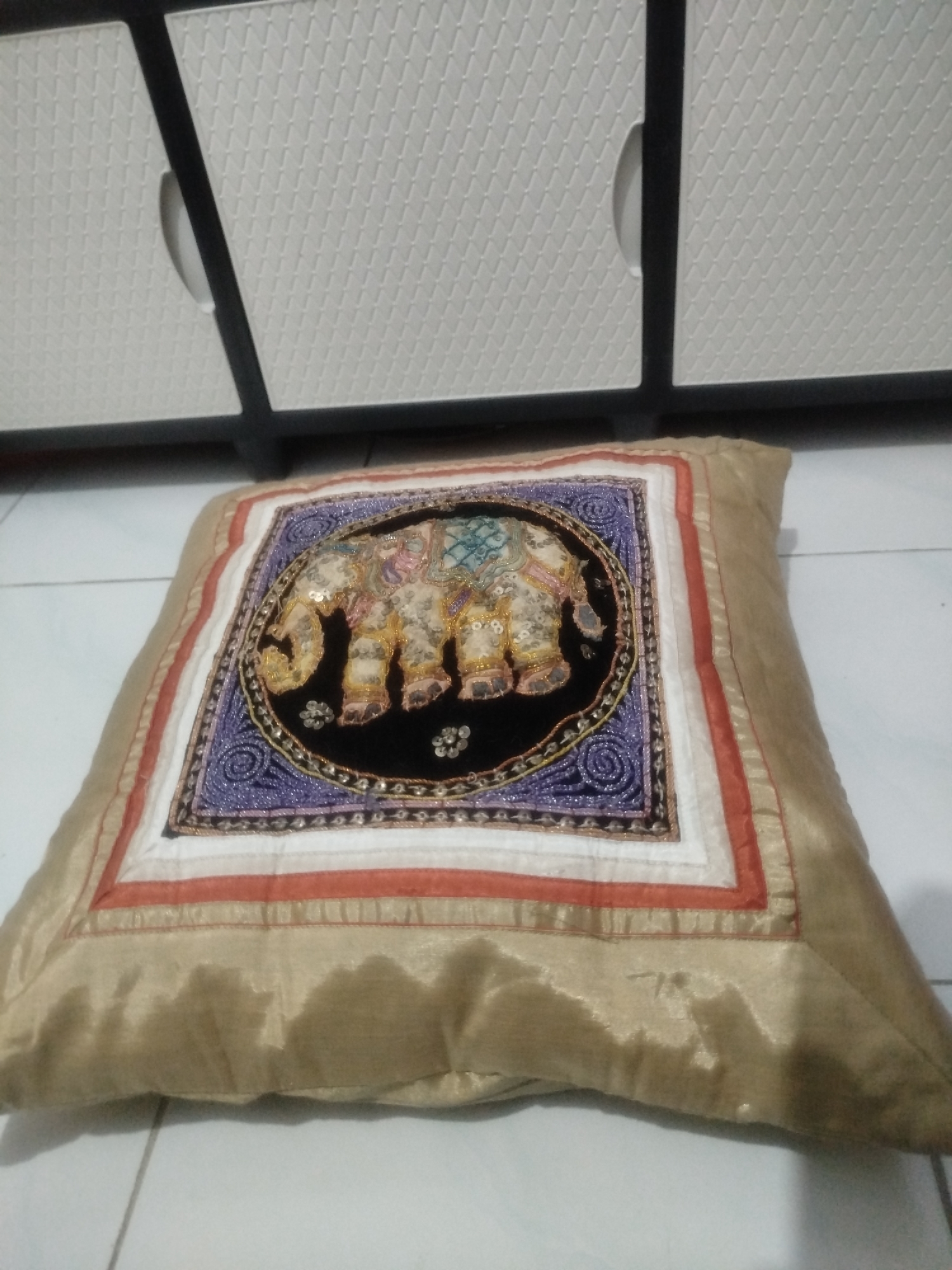 A close-up shot focuses on a square pillow with an intricate elephant embroidery on its front. The pillow is made of a shiny, champagne-colored fabric. The embroidery depicts an elephant in profile, adorned with sequins and colorful threads, set against a black background. This embroidered section is surrounded by layered borders of purple, white, and orange fabric. The pillow rests on a tiled floor. In the background, the lower portion of a white cabinet with textured doors and dark trim is visible, suggesting an indoor setting. The lighting is somewhat dim, with reflections on the cabinet and the pillow's fabric. The scene appears to be in a residential environment within the city of Depok, Indonesia, likely in a living room or bedroom.