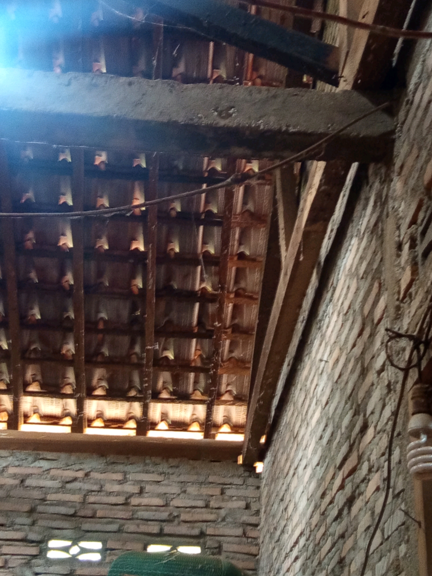 The image depicts the underside of a roof structure, likely in an unfinished or rustic building, possibly located in Karang Sari, Indonesia. The roof is constructed with traditional terracotta tiles, supported by wooden beams and purlins. The tiles are arranged in overlapping rows, with a visible gap between them allowing light to filter through. In the foreground, a brick wall is visible, constructed with rough, reddish-brown bricks. A fluorescent light bulb is mounted on a wire near the wall, suggesting the presence of some electrical installation. The overall impression is of a functional, perhaps agricultural or storage space, characterized by its raw, unrefined construction materials.