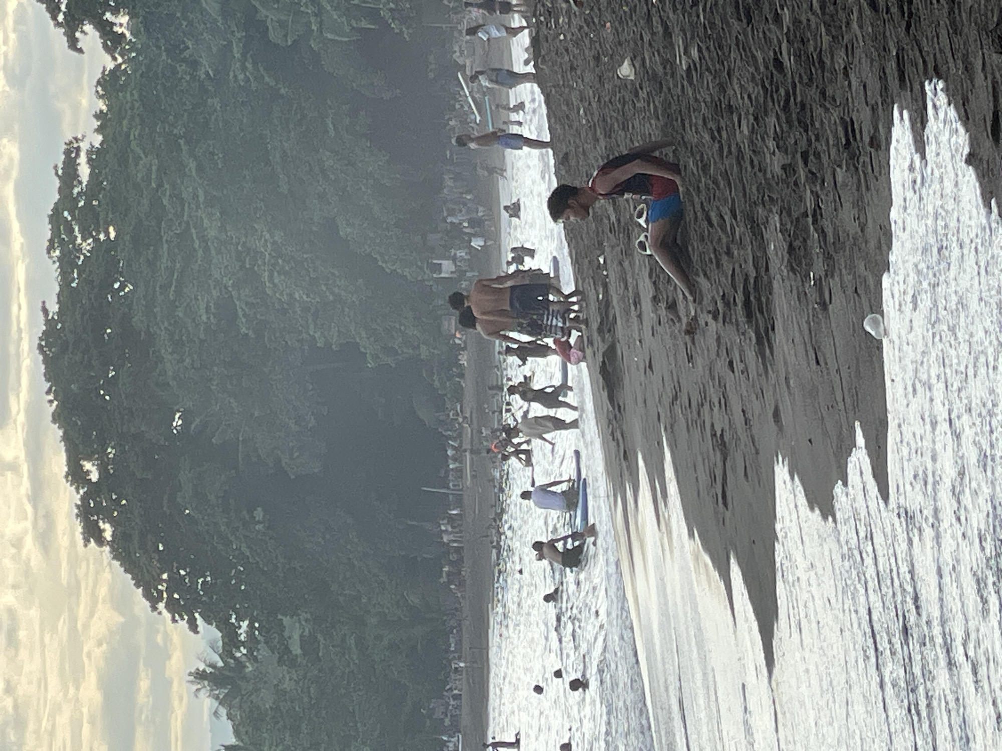 This image depicts a crowded beach in Dauin, Philippines. The foreground features dark, wet sand with white foamy waves lapping at the shore. A young man in red and blue swim trunks is sitting on the sand, facing left, with a pair of flip-flops beside him. In the middle ground, numerous people are in the water, some swimming and others on surfboards. The water appears choppy with white caps. Further back, the beach is lined with many more people, some standing and others appearing to be sitting or walking. A dense, dark green forest with palm trees lines the background of the beach. The sky above is overcast with soft, diffused light, suggesting it could be dawn or dusk. The overall scene conveys a lively atmosphere of a popular beach destination.