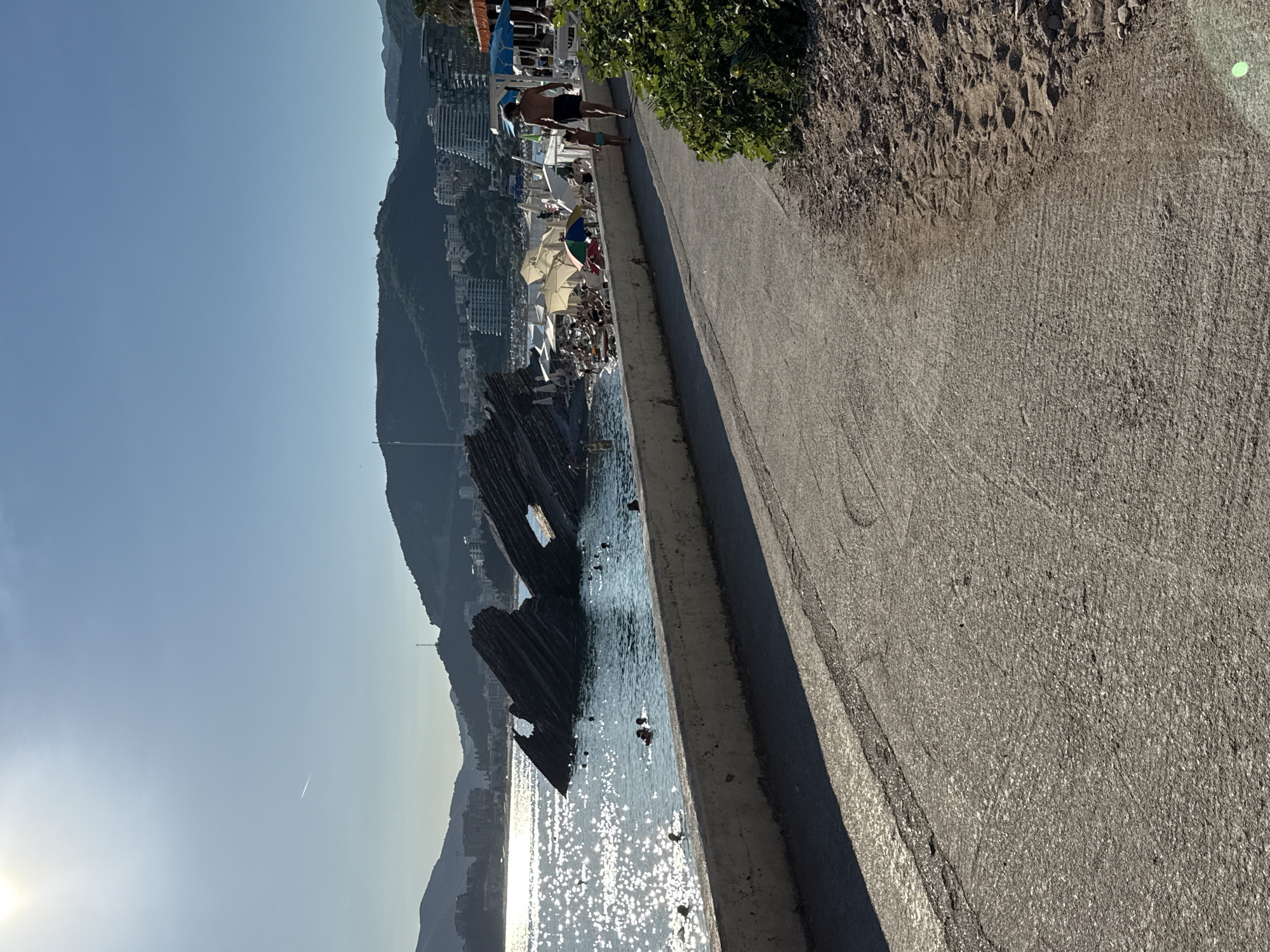 In Rafailovići, Montenegro, a sunlit promenade runs alongside a rocky shoreline. The sea, glinting with sunlight, is dotted with several swimmers. Prominent, angular rock formations emerge from the water, with one featuring a natural arch. Beyond the water, a beach is crowded with numerous people relaxing under umbrellas and lounge chairs. A cluster of white and colorful beach umbrellas are visible, with some structures and buildings lining the shore in the distance. A man and a child walk away from the camera on the promenade, which is bordered by a concrete wall on the left and a gravelly embankment with green foliage on the right. The background consists of lush green hills and a town with buildings and hotels cascading down their slopes. The sky is clear and bright blue.
