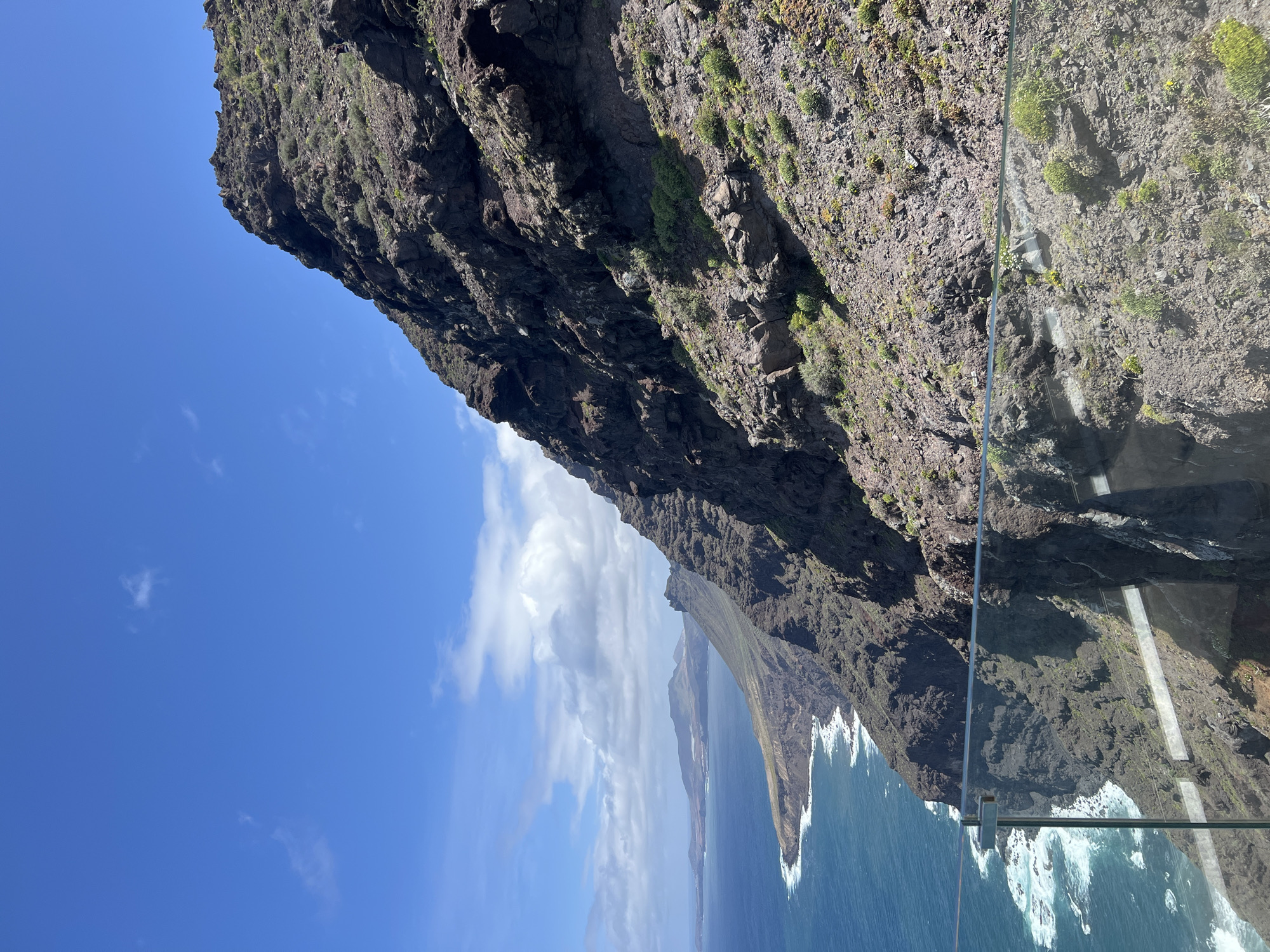 The image shows a view from a glass-floored platform overlooking a cliff and the ocean in Tirma, Spain. The cliff face is rocky, with sparse vegetation. The ocean is blue with white waves crashing against the base of the cliffs. A blue sky with some clouds is visible in the background. The glass platform is visible in the foreground, providing a sense of perspective and depth.