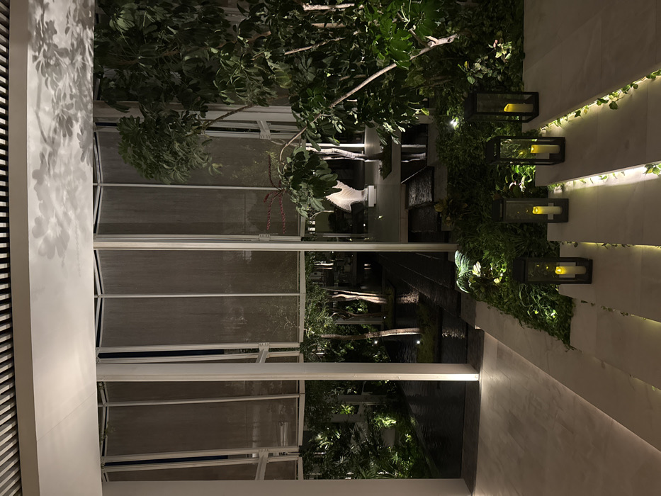 This media file captures an elegant evening scene, likely within a luxury resort or private residence in El Caracol, Mexico. The image, if correctly oriented, shows an interior or covered walkway on the left, leading to an open-air garden or courtyard on the right.
In the foreground and left side, clean white architectural structures are prominent, featuring a white wall with a slatted or paneled section. Distinct shadows of leaves are cast onto this white surface, suggesting light from an unseen source. Large glass panels or windows separate this area from the garden beyond.
The central and right portions of the image showcase a lush, illuminated garden space. Dense green foliage and tree branches fill the view, indicating a tropical or subtropical environment. A notable white, ridged decorative sculpture, possibly abstract or representing a marine creature, stands among the plants. In the mid-ground, a dark, reflective surface, likely a decorative pool or water feature, runs horizontally, mirroring the subtle lights and surrounding greenery.
On the far right, a textured wall is adorned with creeping vines and several contemporary black lanterns, each housing a warm, glowing candle-like light. These lights contribute to the serene and inviting ambiance. The overall impression is one of tranquility and refined design, blending modern architecture with natural elements.
The time of day is clearly night or late evening, as evidenced by the darkness and the reliance on artificial lighting to illuminate the plants and architectural features. No people are visible, and there is no discernible activity taking place. No explicit text is visible in the frame.