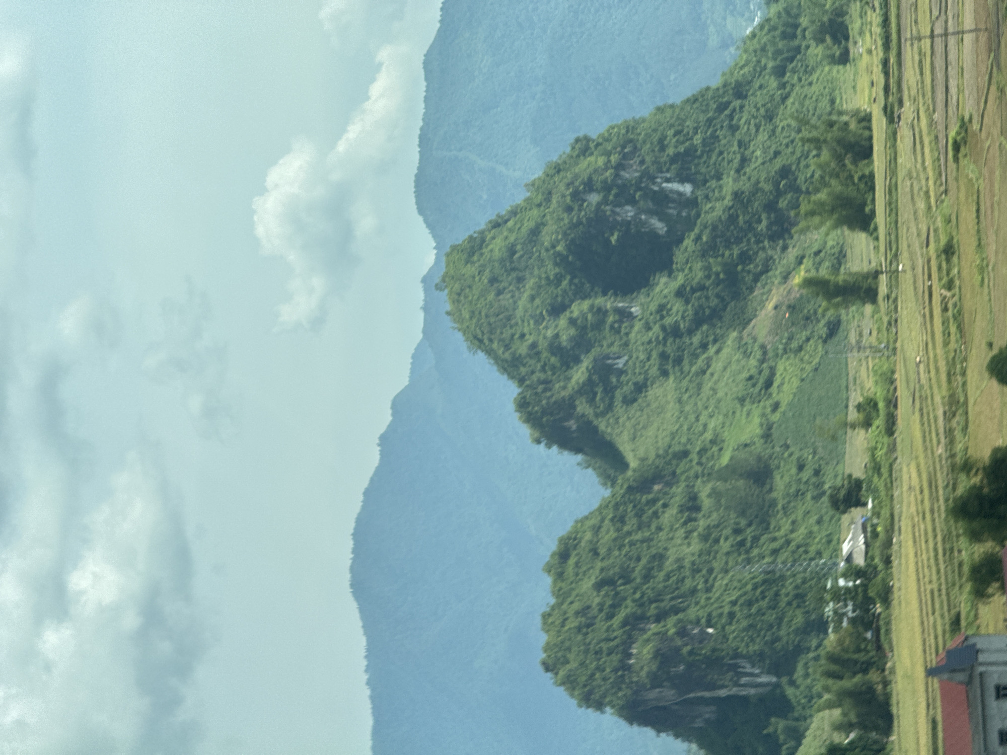 This landscape image, likely taken in Than Uyên, Vietnam, captures a panoramic view of a mountainous region under a partly cloudy sky. The scene is dominated by layers of lush green mountains, characteristic of tropical or subtropical environments.
In the foreground, to the right of the frame (bottom when rotated correctly), there are distinct agricultural fields, possibly rice paddies, laid out in parallel strips of varying green and brown hues, suggesting cultivation. A building with a prominent red roof and grey walls is visible in the bottom right corner of the field area, indicating human settlement. Scattered trees and sparse vegetation also line the edge of these fields, and what appear to be utility poles or power lines can be seen running through them.
The central and left portions of the image feature dense, tree-covered mountains. A prominent, rugged mountain peak, heavily vegetated with various shades of green, stands closer to the viewer. Behind it, further mountains recede into the distance, appearing softer, hazier, and bluer, illustrating atmospheric perspective. The sky above is pale blue to light grey, dotted with fluffy white clouds, suggesting a daytime shot, possibly mid-morning or afternoon, with soft, diffused lighting.
No specific human activity is visible, and there are no people or discernible text in the image. The overall impression is one of a serene, natural landscape interspersed with rural agricultural land.