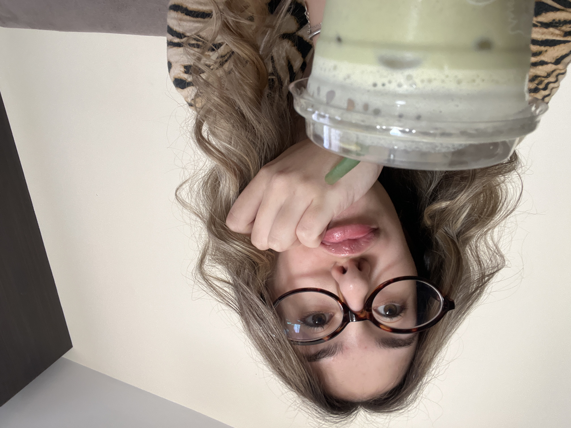 A woman with long, wavy blonde hair and tortoise-shell glasses is holding a clear plastic cup filled with a light green beverage with ice and foam close to her face. She is wearing a patterned top with a dark and light striped pattern. Her lips are slightly parted. The background is a plain off-white wall. There is a dark rectangular object in the lower left corner of the frame. The photograph is taken from a low angle, looking up at the subject, and is oriented upside down. The location is not identifiable.