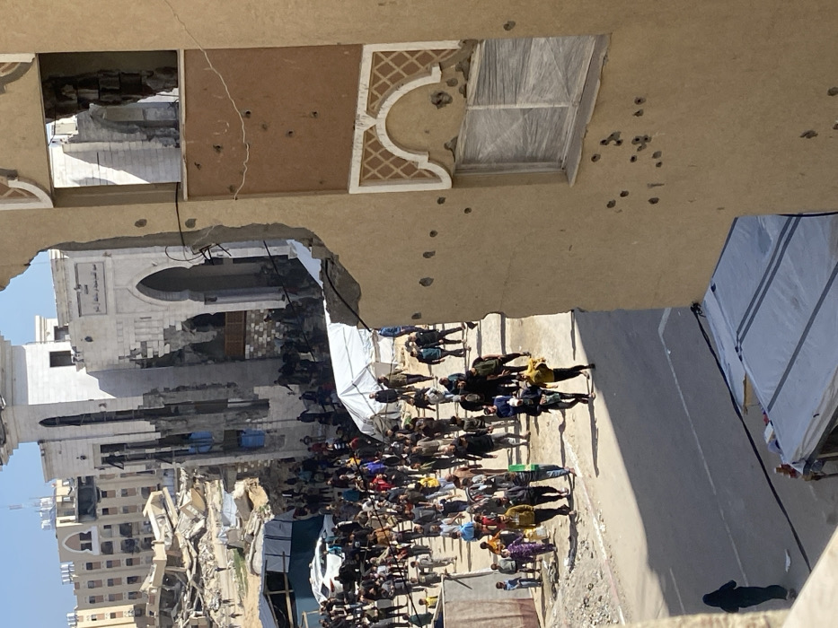 The image depicts a scene of extensive urban destruction in Khan Yunis, Palestinian Territory, under a clear blue sky.
In the foreground, the right side is dominated by a severely damaged multi-story building constructed of light brown/tan material. Its facade features a large, irregular breach revealing interior structural elements. Numerous small, circular impact marks, consistent with bullet holes, are visible across the exterior wall. A window below a decorative arched pattern appears taped or covered.
The midground features a wide, unpaved street or open area, densely populated by an estimated 100-150 individuals. This crowd comprises adult males, women, and children, generally moving on foot. To the left of this gathering, substantial piles of concrete, rebar, and other structural debris are present, alongside at least two white canvas shelters or tents. A white passenger vehicle is partially visible near these structures. At the bottom left, a single individual in a dark garment walks on the street.
The background is dominated by the heavily damaged Great Mosque of Khan Yunis, identifiable by its distinctive arched entrance and a prominent, partially collapsed tower-like structure, likely a minaret. The mosque exhibits significant structural compromise, with large sections of its walls missing. Beyond the mosque, the cityscape reveals further widespread destruction, with numerous multi-story buildings reduced to rubble or severely damaged, extending to the horizon.