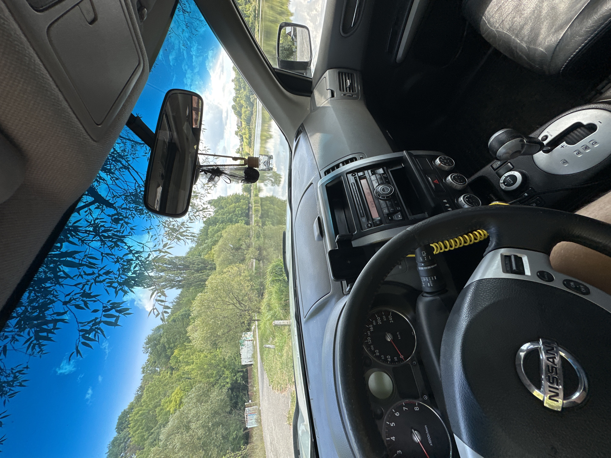 Inside a Nissan X-Trail, the driver's perspective faces forward through the windshield, revealing a picturesque lakeside scene. Lush green trees line the shores of a calm body of water, with a clear blue sky overhead, punctuated by wispy clouds and silhouetted tree branches. The car's interior is visible, including the steering wheel with the Nissan logo, dashboard with gauges and radio, gear shifter, and part of the passenger seat. The location is identified as Makiv, Ukraine, likely on a tranquil summer day.