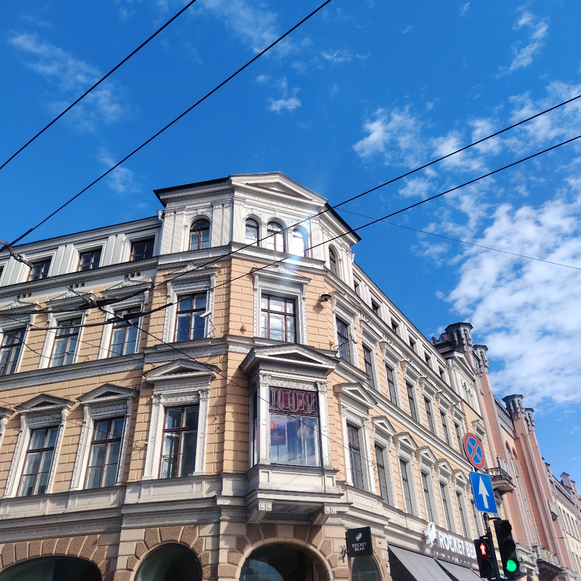 The image shows a street view in Riga, Latvia, looking up at a corner building with an ornate architectural style. The building has a light yellow facade with white window frames and decorative elements. The sky is blue with scattered clouds, and electrical wires are visible running across the top of the frame. A traffic light is showing green and a blue sign with a white arrow points upwards next to a red circle with a blue X. A sign for "Rocket Bean" can be seen at the bottom of the building.