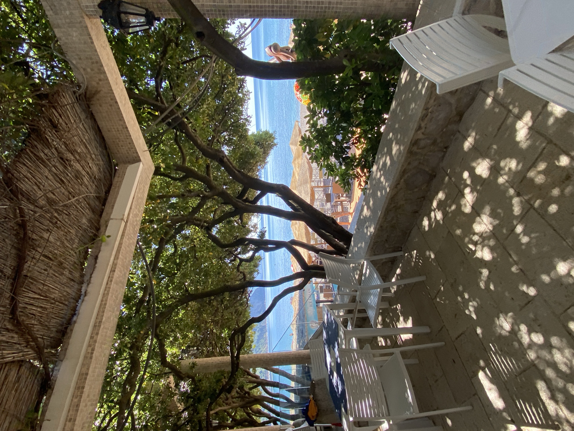 The image depicts a beach scene in Prijevor, Montenegro, viewed from a shaded veranda or covered patio area. The view looks towards the Adriatic Sea, with trees framing the beach. In the distance, there are sunbathers on the beach, along with parasols and chairs. The foreground patio has white plastic chairs and tables, with shadows cast by the trees and sun. The patio is constructed with stone. The image is angled, as if the camera was tilted during the capture. A straw-covered roof structure is visible to the left.
