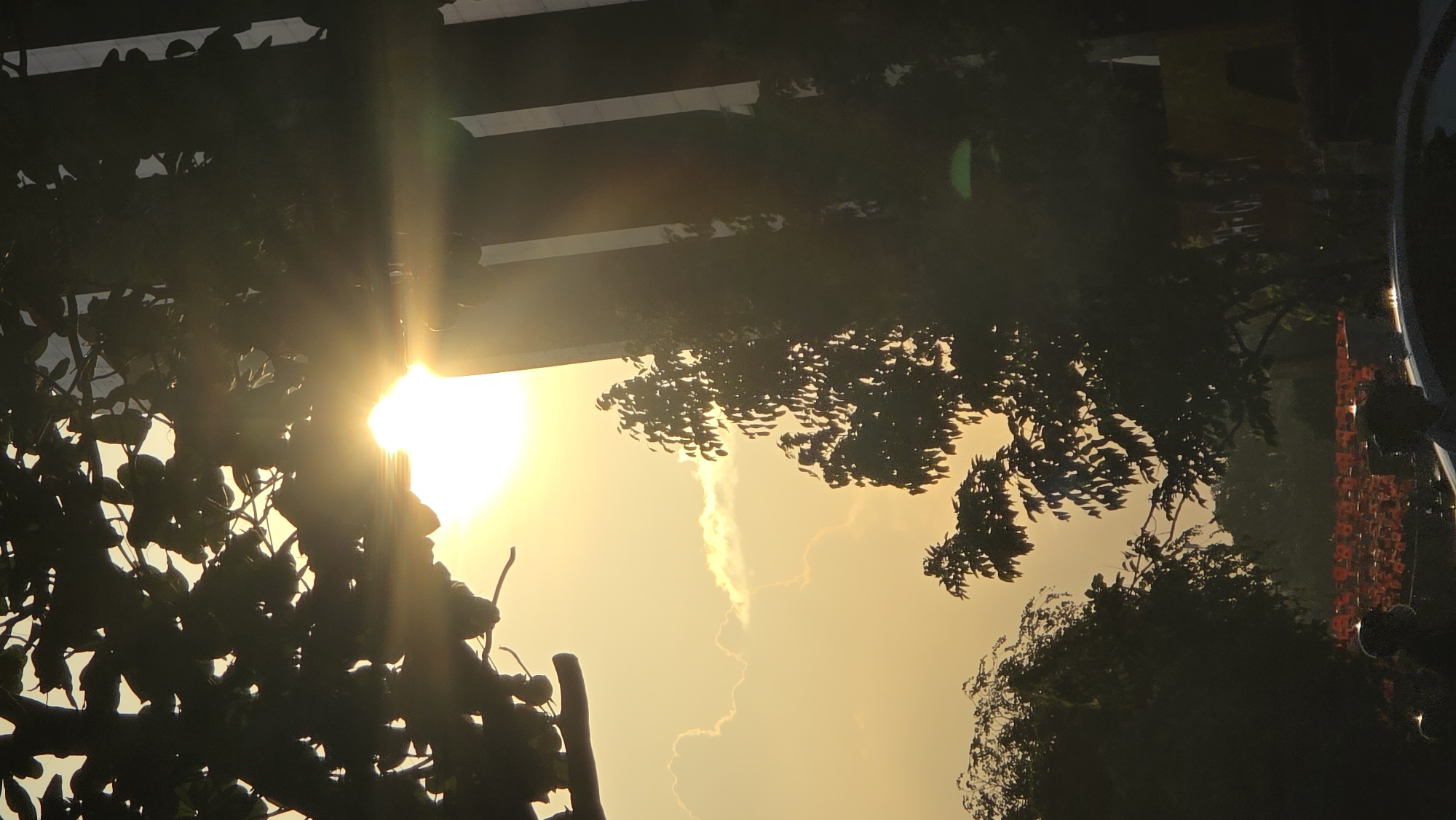 The image captures a sunlit scene in Nha Trang Ward, Vietnam. The sun, positioned near the center, casts a bright glow through foliage and the lower section of a building's structure. Silhouetted trees and leaves frame the sunlight. A wisp of cloud is visible in the sky. In the background, parts of a building, including what appear to be decorations or lighting, are discernible. A portion of a vehicle is also visible. The overall environment is bright and warm.