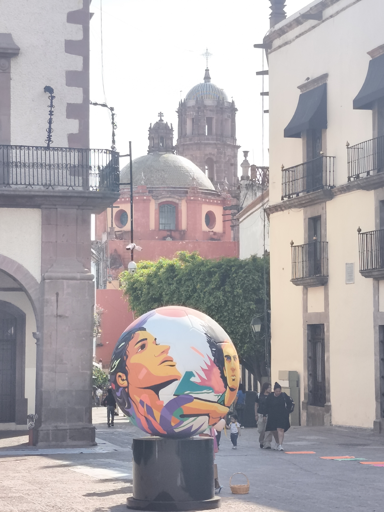 The image shows a plaza in Querétaro, Mexico, with a large painted soccer ball sculpture on a black pedestal in the foreground. The ball features stylized faces, painted in bright colors. Behind the sculpture, there is a view through a stone archway, revealing a church with a large dome and a tower topped with a cross. The buildings surrounding the plaza are light-colored, with dark awnings and iron balconies. Several people are walking in the plaza, including a woman in a black dress and a child holding her hand. There are colorful mats lying on the ground to the right. The sky is bright, suggesting it is daytime.