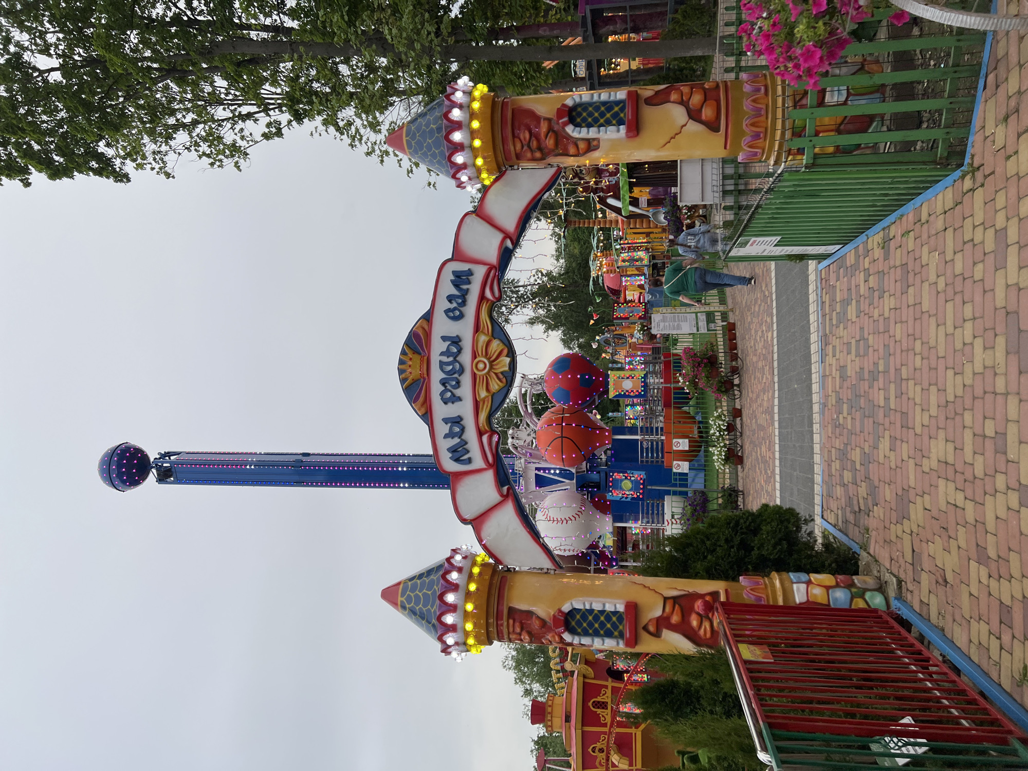 The image shows an amusement park entrance in Mytishchi, Russia. A large archway welcomes visitors. The sign on the archway is written in Russian. Decorations include castle-themed towers, sports balls (basketball, baseball), and flowers. A tall ride with a ball-shaped end extends high into the cloudy sky. A paved walkway leads into the park. A person wearing a green shirt and jeans is walking along the walkway. Green metal fencing borders the walkway.