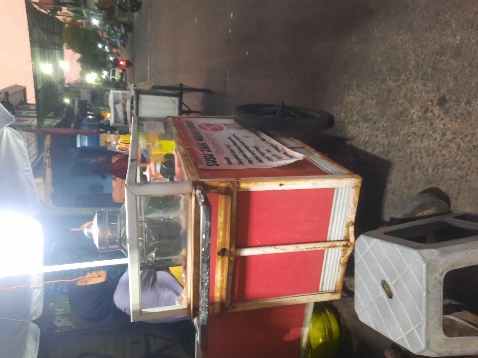 An outdoor, low-angle, slightly blurry, medium shot shows a food cart on a street in North Jakarta, Indonesia. The cart is red with white trim and a glass display case on top. A woman wearing a dark hijab and a purple shirt is visible behind the cart, appearing to be preparing food. Various containers, bottles, and utensils are on display on the cart. A sign on the side of the cart reads "SUSU JAHE MERAH SU." The cart is parked on a paved street with a curb and a sidewalk. In the background, there are other food carts, buildings, and a few motorcycles. The sky is a dusky orange, suggesting it is either dawn or dusk. A white plastic stool is in the foreground.