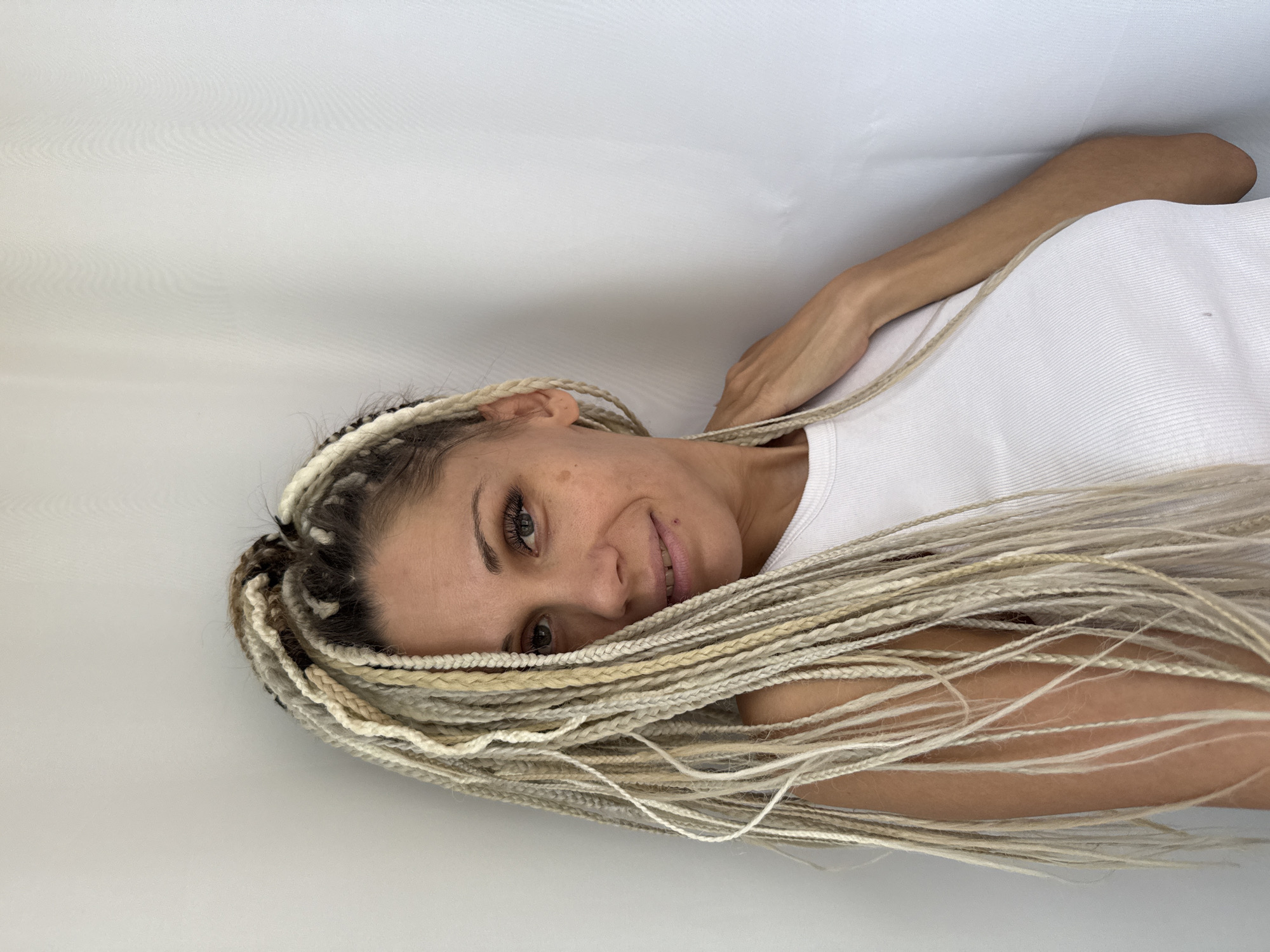 A woman with long, blonde braids stands leaning against a white wall in what appears to be an indoor setting. She is wearing a white ribbed tank top. Her gaze is directed towards the camera with a slight smile. The lighting is soft and diffused, casting no harsh shadows. The background is plain and uncluttered, with the white wall occupying the majority of the frame. There is no visible indication of the city of Rusanovo, Russia, in the image.