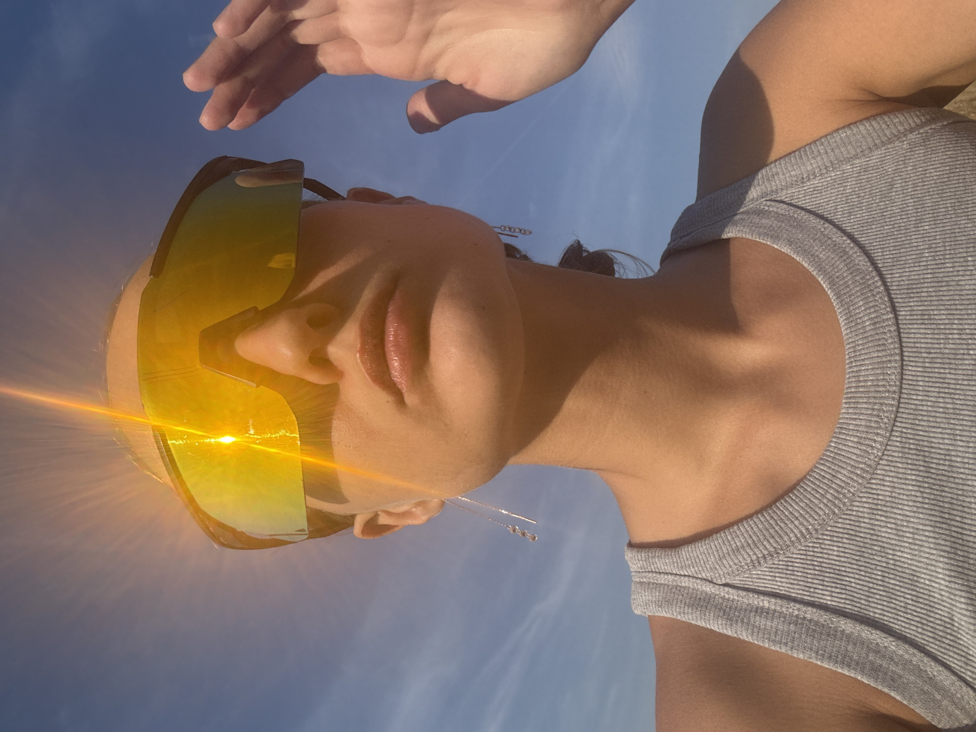A low-angle, close-up outdoor shot shows a person wearing large, reflective yellow sunglasses and a grey ribbed tank top. The sun glares brightly on the glasses, creating lens flare. The person is raising their right hand to shield their eyes. A blue sky with wispy clouds forms the background. The location is identified as Podshivalovo, Russia.