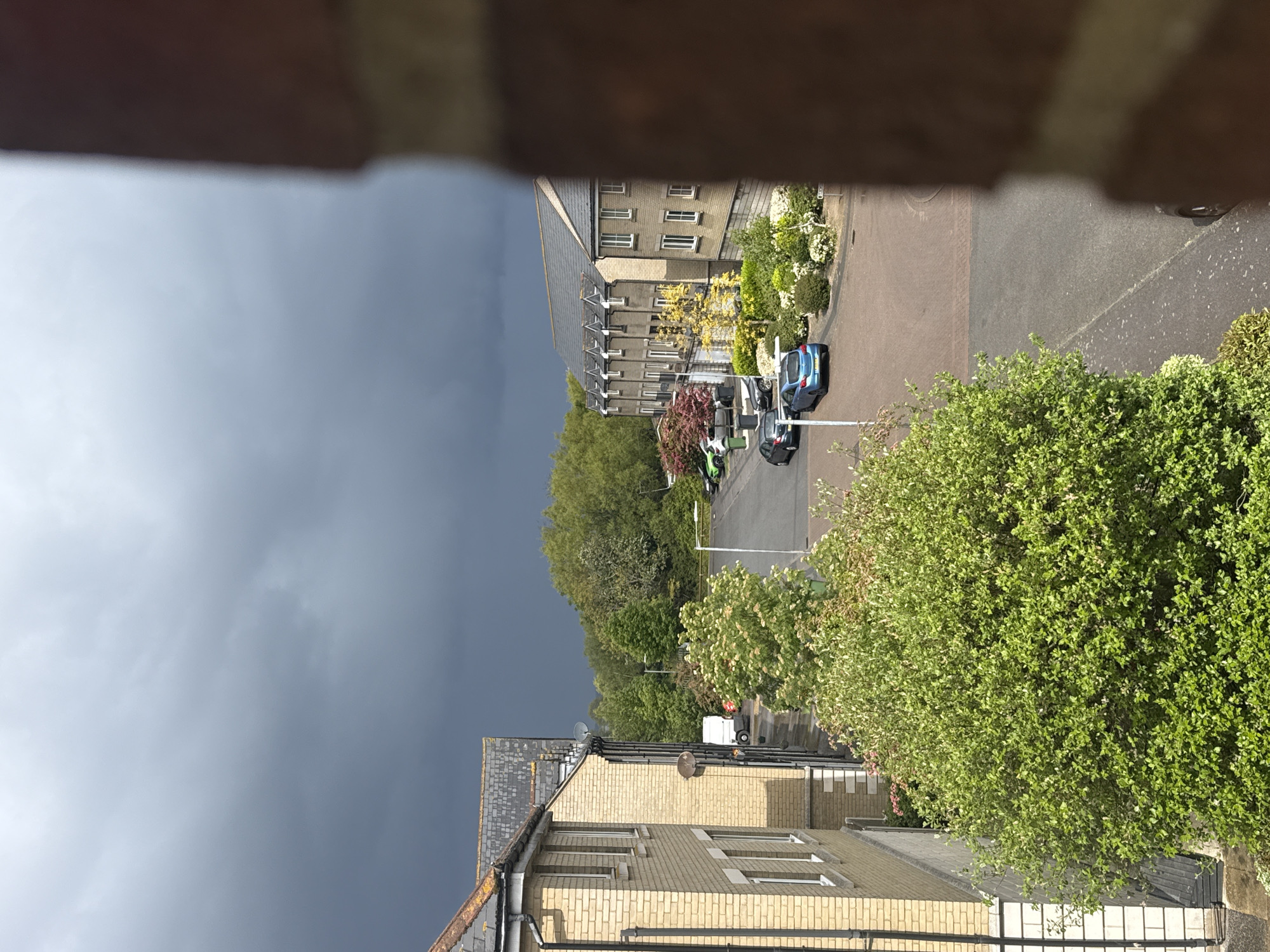 The image depicts an elevated view of a street scene in Great Yarmouth, United Kingdom. Buildings constructed of light-colored brick with dark tiled roofs are visible, interspersed with mature trees and bushes. Dark storm clouds fill the sky. A street with parked cars runs through the scene. No people are visible. The photo appears to be taken from an upper floor of a building, as evidenced by the obscured view at the top.