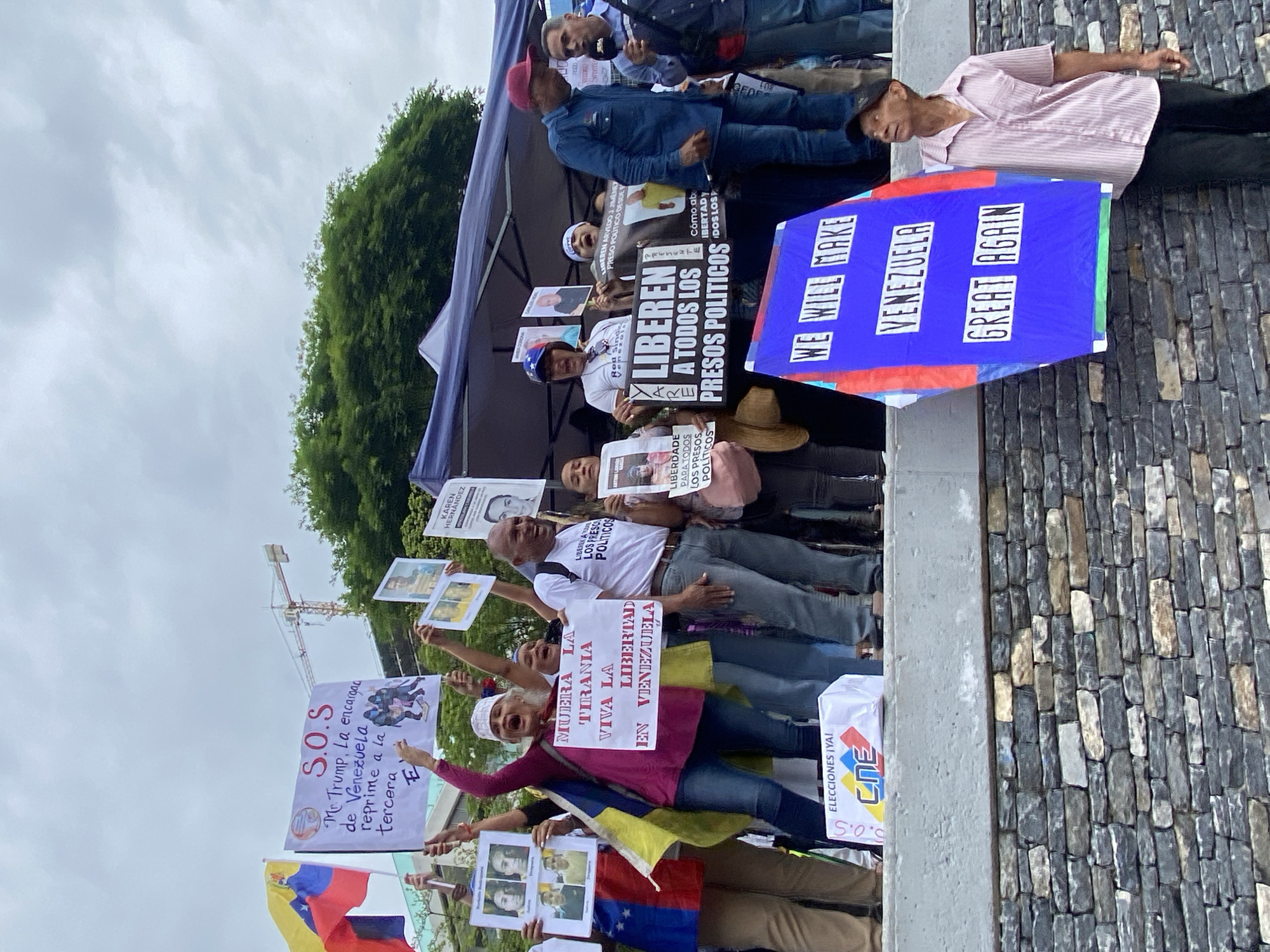 This image captures an outdoor political demonstration in Caracas, Venezuela, under an overcast sky during the daytime. A diverse group of men and women are gathered, holding various signs and Venezuelan flags, conveying a strong sense of protest and advocacy for freedom and political change.
In the foreground and mid-ground, multiple individuals are visible. Many are holding white placards with hand-written or printed messages. One prominent sign, held by a woman in a magenta shirt and white head covering, reads "MUERA LA TIRANIA VIVA LA LIBERTAD EN VENEZUELA" (Death to tyranny, Long live freedom in Venezuela). Another significant sign, centrally located, states "LIBEREN A TODOS LOS PRESOS POLITICOS" (Free all political prisoners). Several smaller signs feature photographs of individuals, likely political prisoners, with names like "KAREN HERNÁNDEZ" and messages such as "LIBEREN ARVEDO Y JIMENEZ PRESO POLÍTICO DESDE F". A large blue sign with white lettering declares "WE WILL MAKE VENEZUELA GREAT AGAIN." Other visible text includes "LIBERTAD PARA TODOS LOS PRESOS POLÍTICOS," "S.O.S Mr. Trump, La encargada de Venezuela reprime a la tercera E," and "ELECCIONES ¡YA! CNE S.O.S."
The demonstrators appear passionate, with some exhibiting expressions of shouting or chanting. They are dressed in casual attire, including jeans, t-shirts, and button-down shirts. In the background, a large tent provides shade, and a stone-patterned wall borders the area. A construction crane is faintly visible in the distance under the cloudy sky, suggesting an urban setting. The overall mood is one of serious determination and a collective call for political reform and human rights in Venezuela.