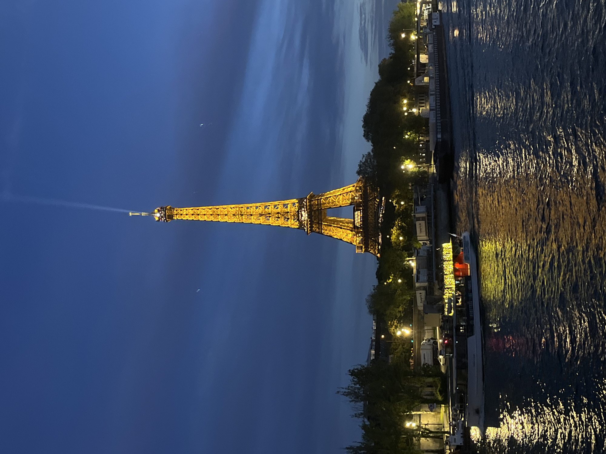 The image depicts the Eiffel Tower illuminated at night in Paris, France. The tower is golden and stands tall against a dark blue sky with some wispy clouds. A beam of light projects upwards from the tower's peak. To the right of the tower is the Seine River, with light reflecting on the water's surface. Along the riverbank, there are trees and buildings with visible lights. A tour boat is docked along the Seine.