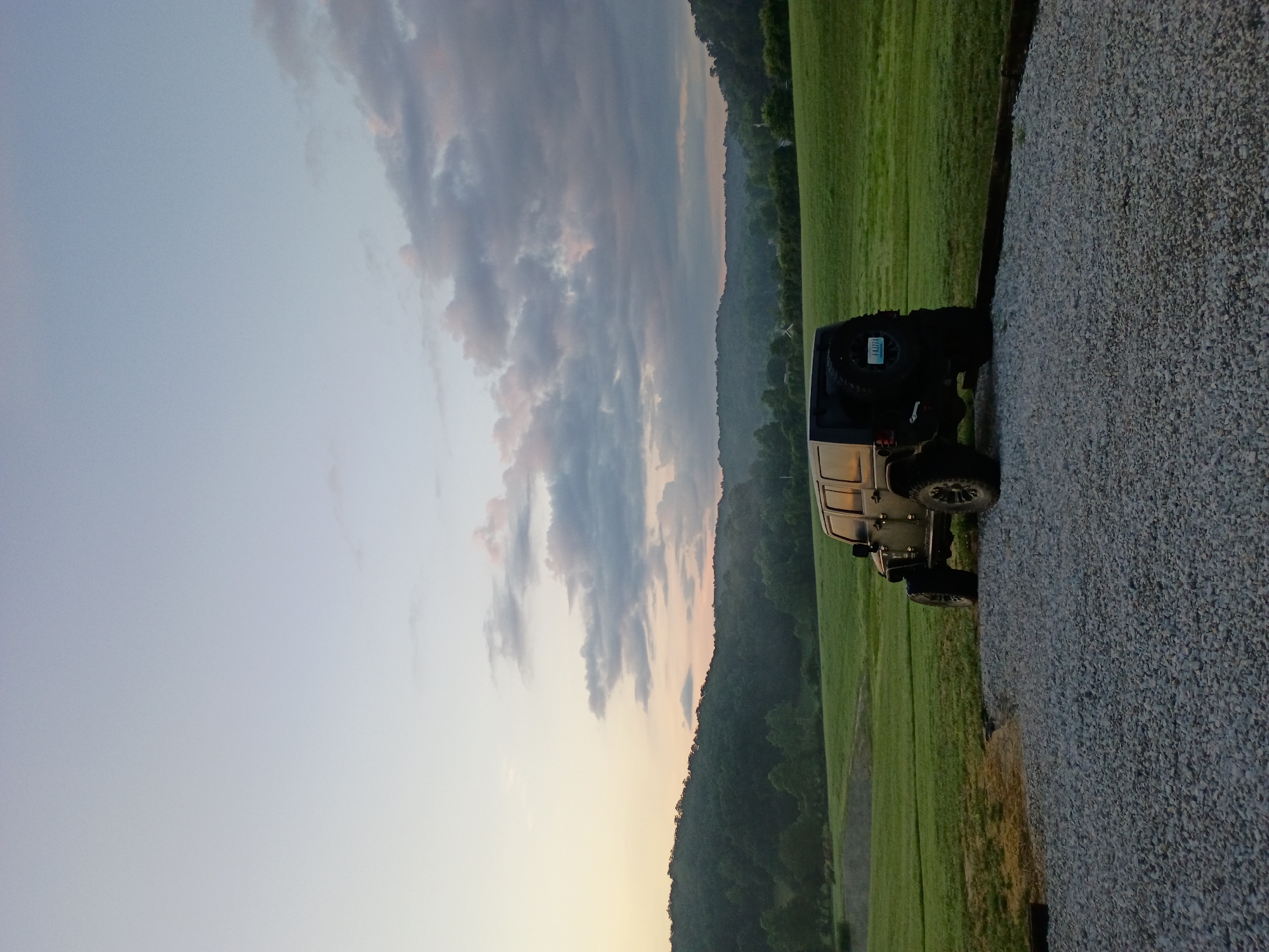 A black Jeep Wrangler is parked on a gravel driveway, facing away from the camera and slightly to the right. The vehicle is positioned in the foreground, with its rear spare tire visible. The background consists of a vast, gently rolling green field that extends towards a tree-covered hillside. The sky above is a soft gradient of light blue and pale orange, with scattered, wispy clouds, suggesting either sunrise or sunset. The scene is set in a rural environment, likely in or around Jeffersonville, United States of America. No people are present.