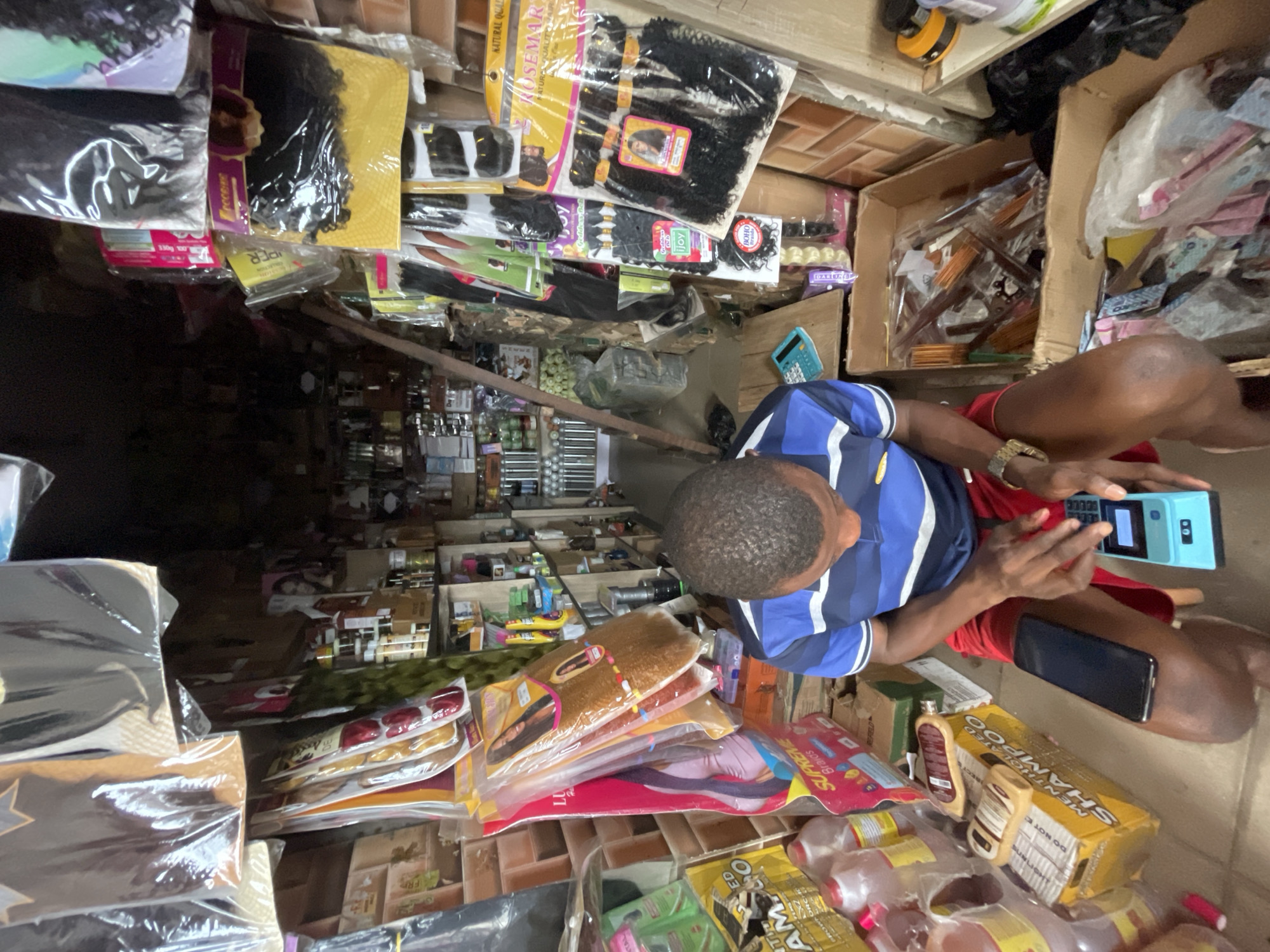 Here is a description of the image:
**Environment:** The image shows a densely packed retail space, likely a small shop or stall. The location is identified as Lekki, Nigeria. The space is filled with a variety of hair products including packaged hair extensions, shampoos, and possibly other beauty supplies. The goods are stacked high and appear to be in a somewhat disorganized manner, filling the frame from floor to ceiling.
**Subjects:** The primary subject is an individual, seen from above, sitting in the space. The person is wearing a blue and white striped shirt and red shorts. They are holding a light-blue mobile device with a calculator user interface, possibly a point-of-sale system or mobile point-of-sale (mPOS).
**Objects:** Various hair products are prominent, including packaged hair extensions of different colors and styles, shampoo bottles and boxes. A black mobile phone can be seen on the person's leg.
**Actions/Interactions:** The person is interacting with the mobile device, suggesting they may be managing sales, inventory, or other business-related tasks.