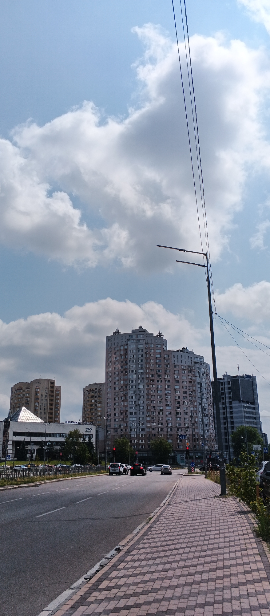 This is a vertical, eye-level, outdoor photograph taken on a partly cloudy day in Kyiv, Ukraine. The foreground shows a paved sidewalk with a pattern of red and grey rectangular pavers. Beyond the sidewalk is a multi-lane road with a metal barrier separating the traffic lanes from a grassy verge. Several cars are driving on the road.
In the mid-ground, a row of trees lines the road, partially obscuring the lower floors of several large, multi-story buildings. The most prominent building is a tall, light-colored apartment complex with numerous windows and decorative elements on its roofline. To its left is a triangular-shaped building with a glass facade, and to its right is a darker, more rectangular skyscraper.
The background consists of a blue sky with scattered white and grey clouds. Overhead, several black electrical wires are visible, suspended from a tall, dark pole that also supports two streetlights. The overall scene depicts a street view in a modern urban environment.
