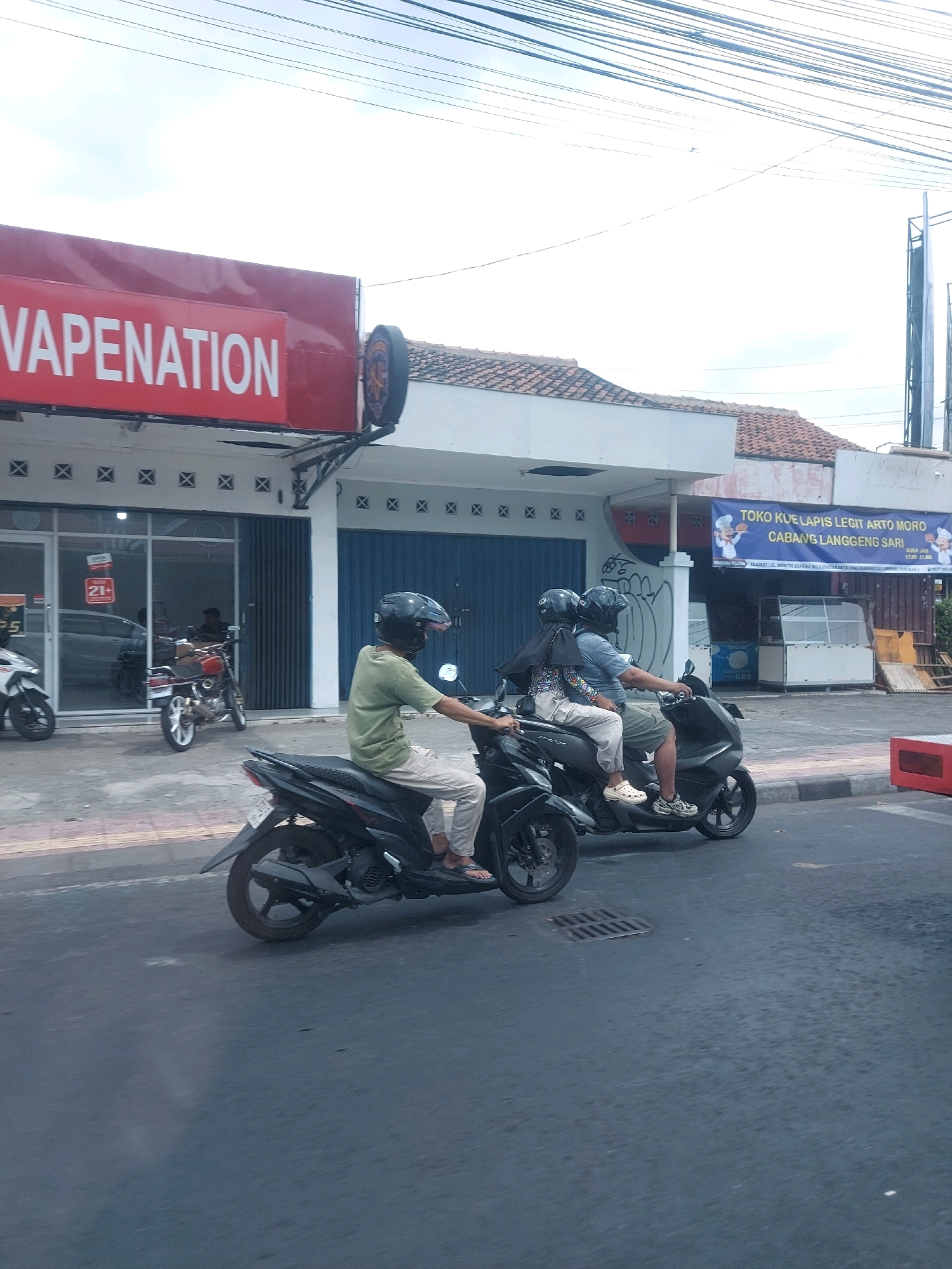 The image shows a street scene in Yogyakarta, Indonesia, with two motorbikes in the foreground. The motorbike on the left is partially visible, showing its rear wheel and a person's legs wearing light-colored pants and sandals. The motorbike on the right is more fully captured, with two people riding it. The rider in front is wearing a green t-shirt, light-colored pants, and sandals. The passenger behind is wearing a patterned shirt and light-colored pants. Both riders are wearing helmets.
Behind the motorbikes, there are buildings. On the left, a shop with a red sign that says "VAPENATION" is visible. The shop has large glass windows and a metal roll-up door. A red motorbike is parked in front of the shop.
On the right side of the image, a banner for "TOKO KUE LAPIS LEGIT ARTO MORO CABANG LANGGENG SARI" is hanging above a shop. The banner has images of chefs and text in Indonesian. The shop appears to be a bakery with display cases. The street in front of the shops is paved, and there are power lines overhead. The overall impression is a bustling urban environment in Yogyakarta.