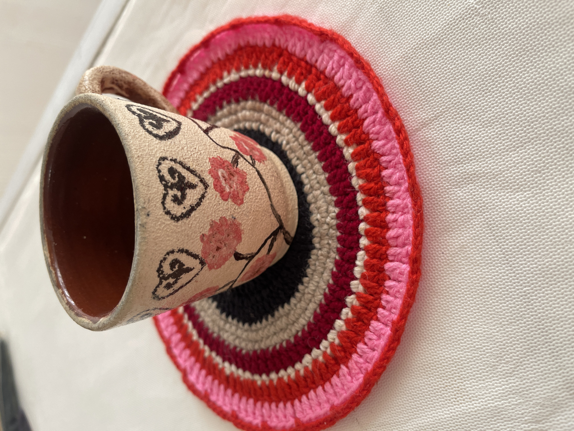 A close-up shot shows a handcrafted ceramic cup with a light beige exterior, decorated with stylized black hearts and pink floral patterns, resting on a crocheted coaster. The coaster is circular and features concentric rings of various shades of pink, red, and black, with a beige outermost ring. The cup has a brown interior and a visible handle on the right side. The items are placed on a textured white surface, likely a tablecloth, with a shallow depth of field blurring the background. No people or explicit location details are visible in the image.