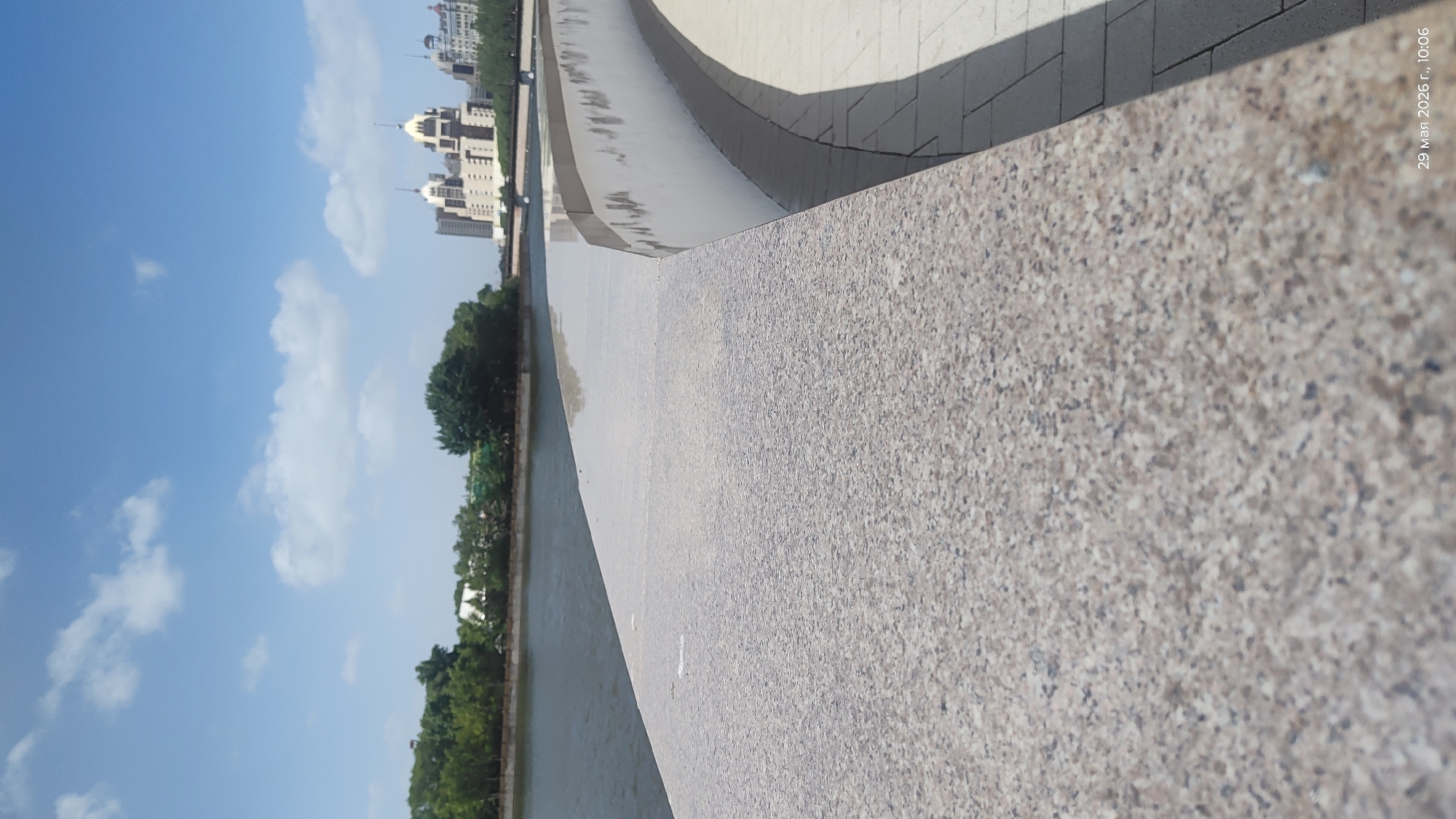 The image presents a view from a bridge or promenade, possibly along a river or canal in Astana, Kazakhstan, as suggested by the architectural style of the buildings in the background. The photograph is taken from a low angle, emphasizing the granite or concrete railing of the structure in the foreground. The railing is a light grey color with a speckled texture. Beyond the railing, there's water reflecting the sky and adjacent greenery. Trees line the bank opposite the viewpoint. In the background, there are buildings with elaborate designs, typical of modern architecture found in Astana. The sky is blue with scattered clouds. A timestamp in the top right corner reads "29 Mar 2026 г., 10:06".