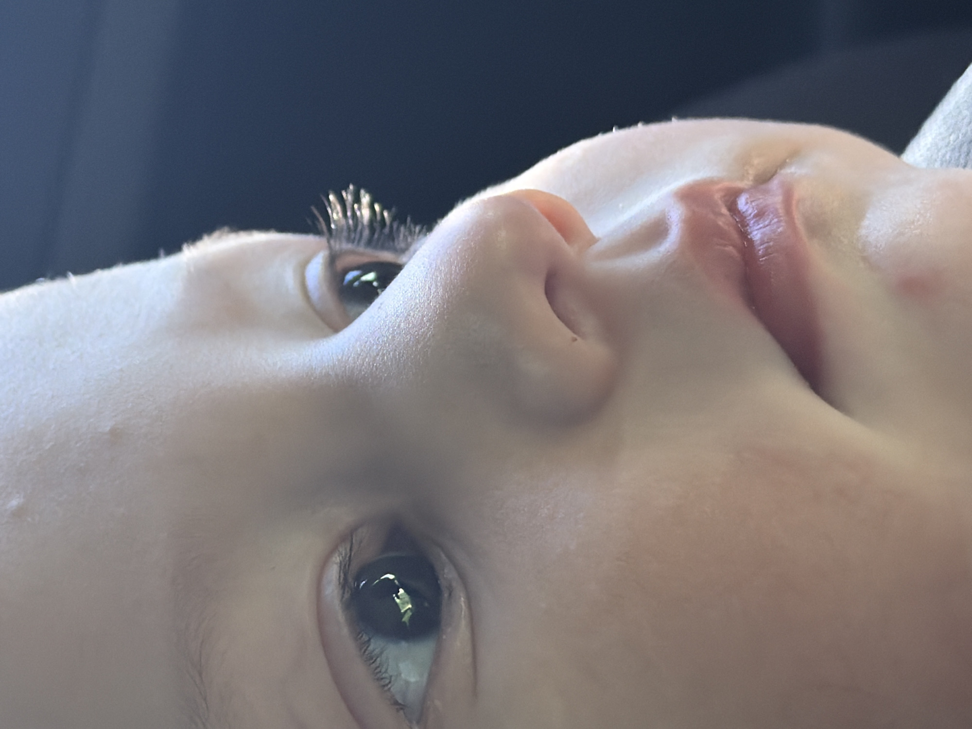 A close-up, side profile photograph shows the left side of a young child's face. The child's left eye, with long eyelashes, is visible, as is their nose and lips. The background is dark and out of focus. The image was taken in Chaikovsky, Russia.