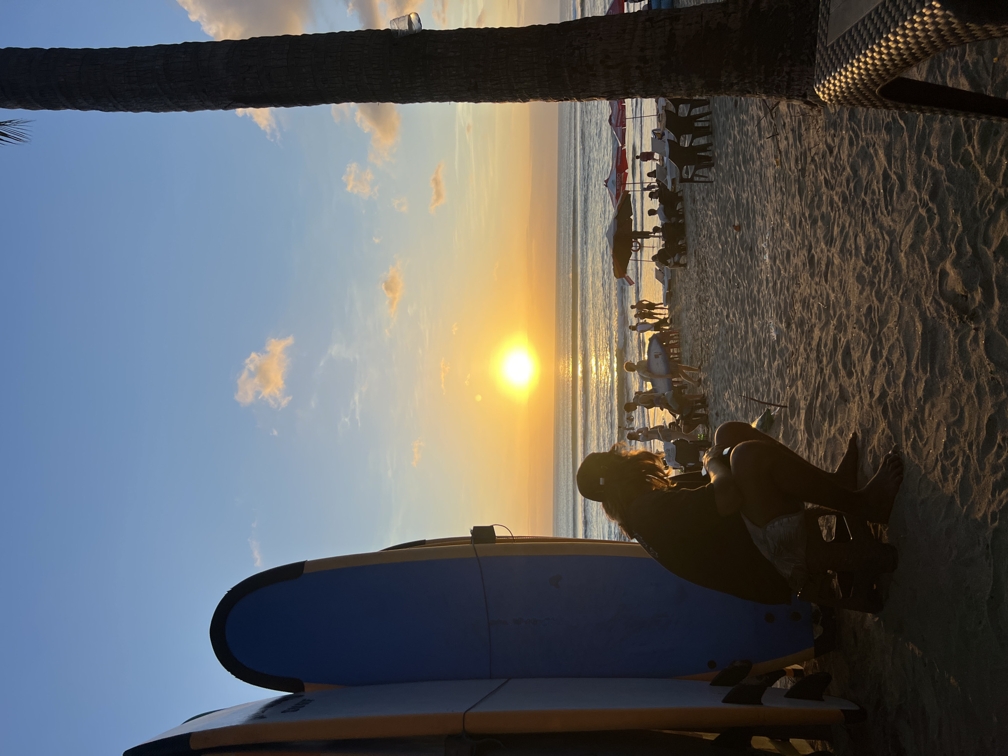 The image depicts a beach scene at sunset in Kuta, Indonesia. In the foreground, a person is seated on a low stool next to several surfboards, facing away from the camera. The person is wearing a dark shirt and shorts, and a cap. The surfboards are tall and appear to be arranged upright.
The background shows the ocean with gentle waves, and the sun is setting on the horizon, casting a warm glow across the water and the sky. Several people are visible on the beach, some standing and others seated under umbrellas. There are lounge chairs and tables set up on the sand. A palm tree trunk is prominently positioned on the right side of the frame, partially obscuring the view. The sand is textured with footprints and shadows. The overall atmosphere is tranquil and evokes a sense of a late afternoon on a tropical beach.
