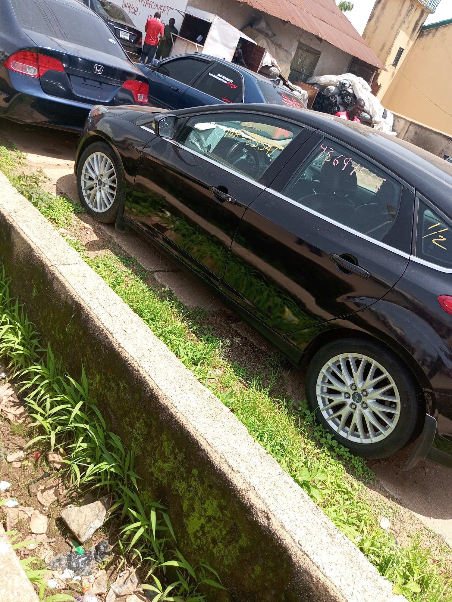 The image shows an outdoor scene with several cars parked. In the foreground, a black sedan is parked next to a concrete barrier overgrown with grass. The car has its doors closed and its tires are visible. In the background, a dark blue sedan is parked behind the black one. Further back, a man in a red shirt is interacting with another person dressed in green, near a building with a corrugated metal roof. There are also large bundles of what appears to be some sort of material wrapped in white plastic. The overall setting appears to be a yard or lot where vehicles are kept, possibly a car lot or repair area, with a building and surrounding vegetation. The ground is dirt and gravel.
Visible text on the black car windows includes "1/2" written in yellow. On the side of the blue car, there is text that appears to read "RECOVERY" and some numbers. The scene is likely set during the daytime, with bright sunlight casting shadows. The location context provided is Bukuru, Nigeria. There are no strong indicators of a specific event or emotion, other than the transactional interaction between the people in the background.